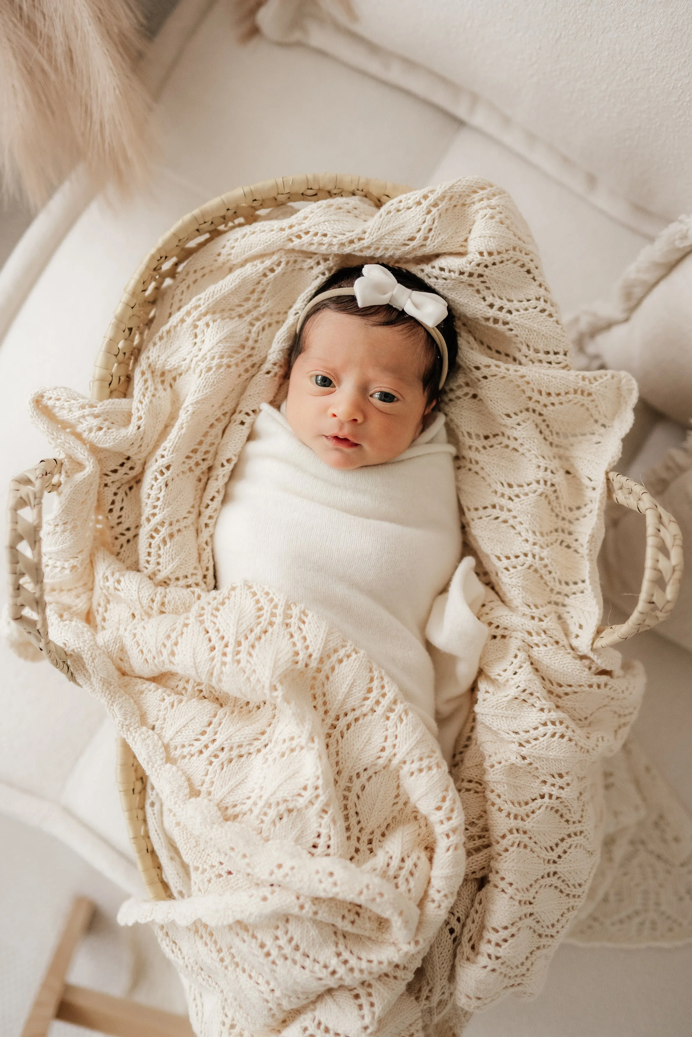 A newborn baby wrapped in white clothing, lying in a woven basket surrounded by cream-colored knitted blankets, with a white bow headband on her head.