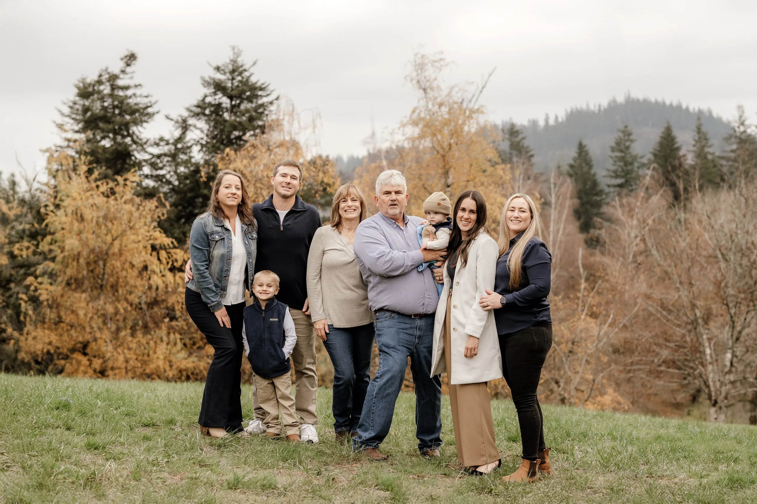 Family photo outdoors during fall, with trees and mountains in the background, showing seven people of various ages smiling and posing together.