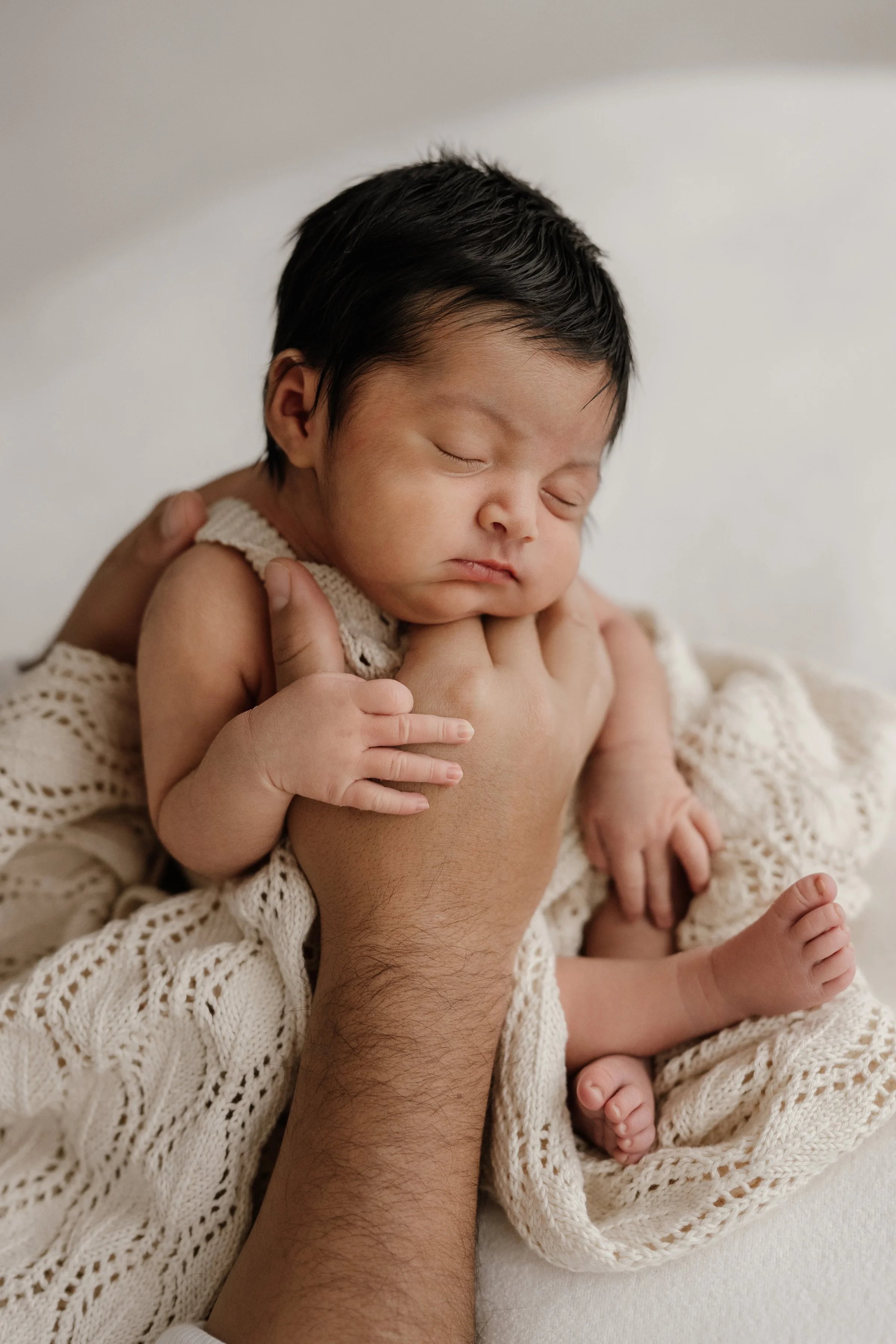 A newborn baby with dark hair sleeping peacefully, being gently held by an adult's hand, wrapped in a creamy knitted blanket.