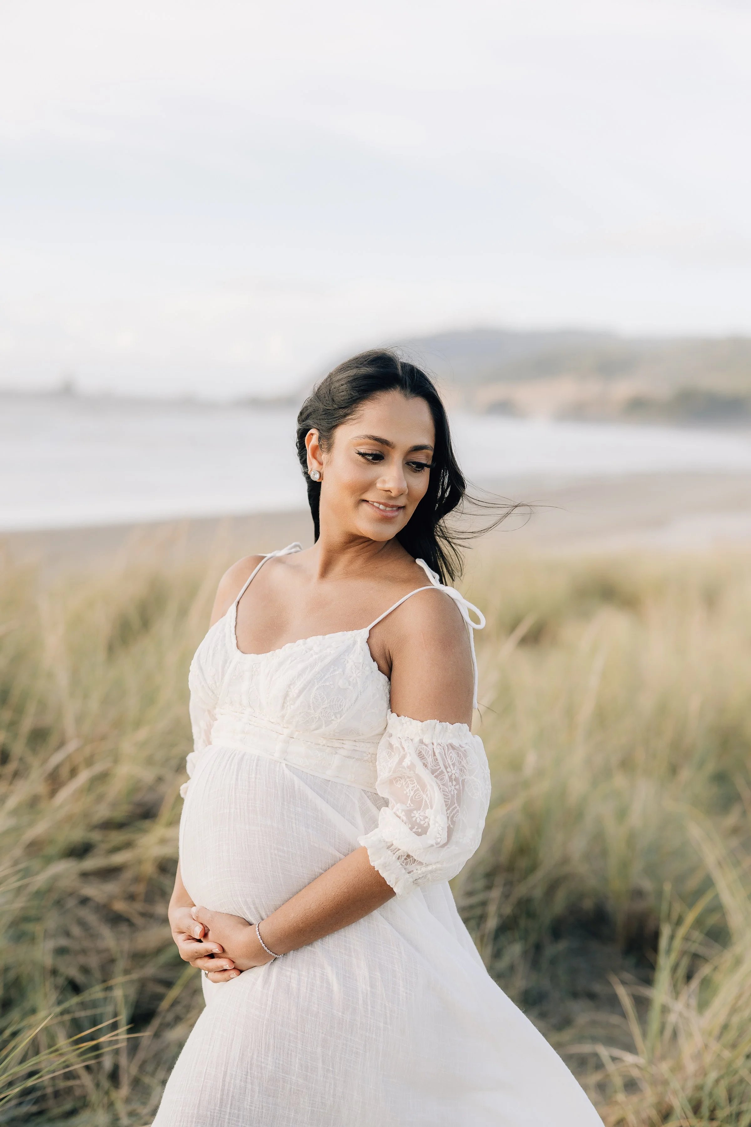 A pregnant woman in a white dress standing outdoors on a beach with grassy dunes and water in the background, looking down and smiling gently.