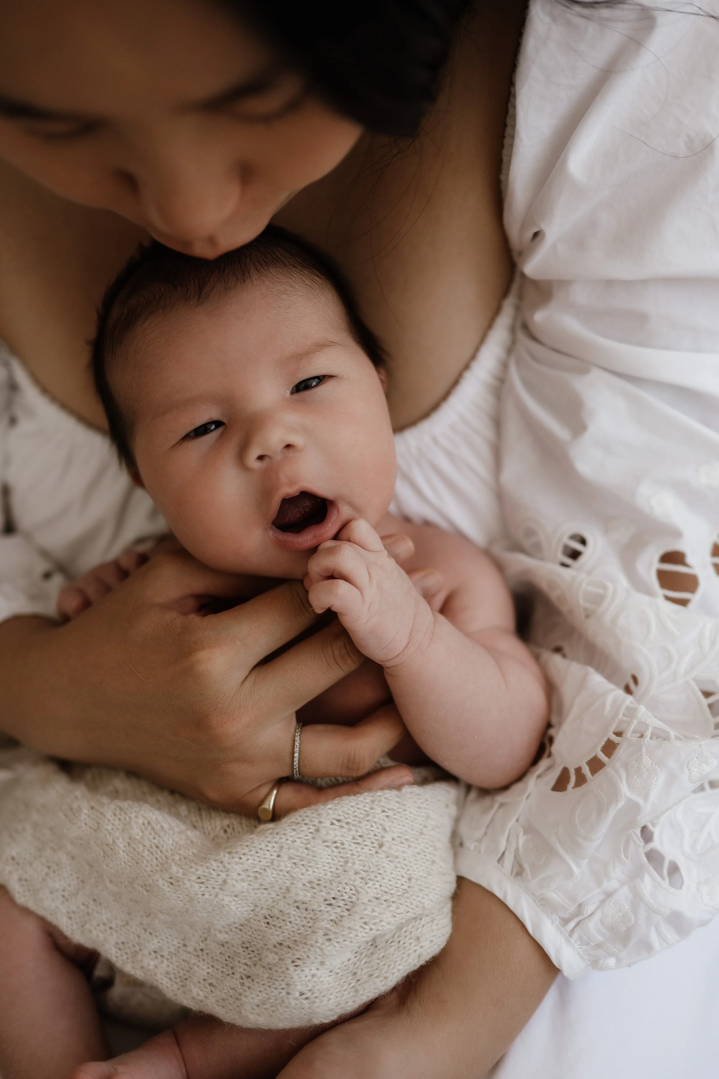 Close-up of a woman holding a yawning baby with open eyes, wearing a white embroidered top, with the woman wearing rings on her fingers.