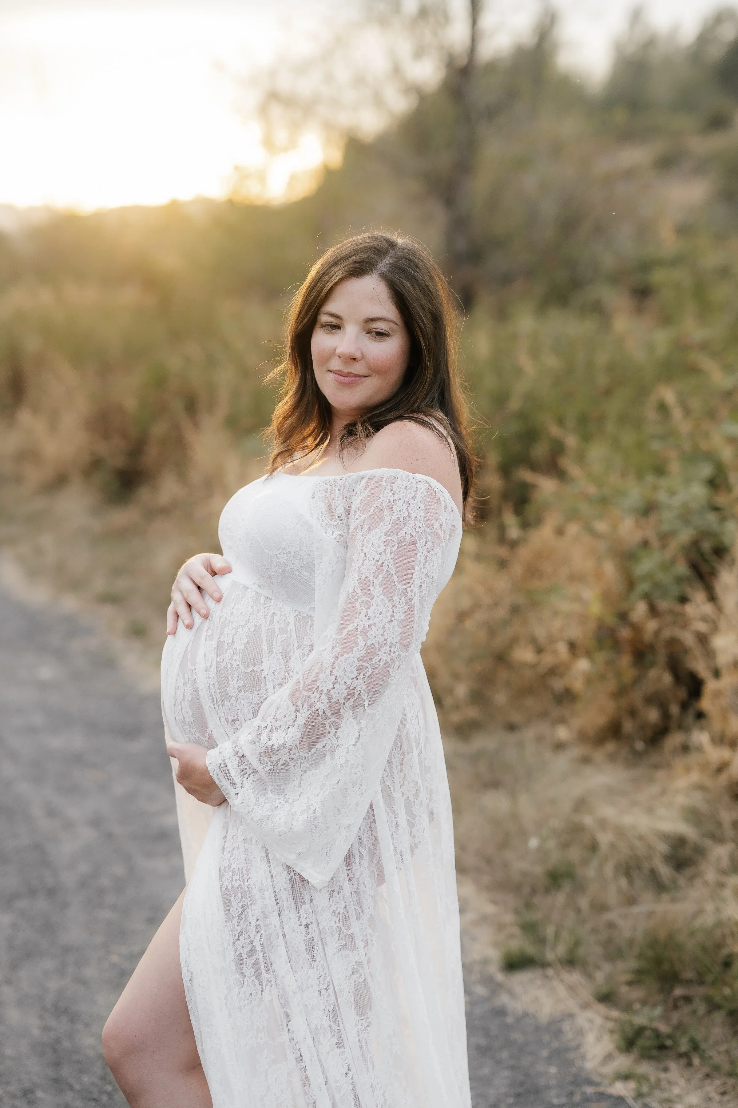 Pregnant woman in a white lace dress standing outdoors during sunset, with her hand on her belly and looking downward.