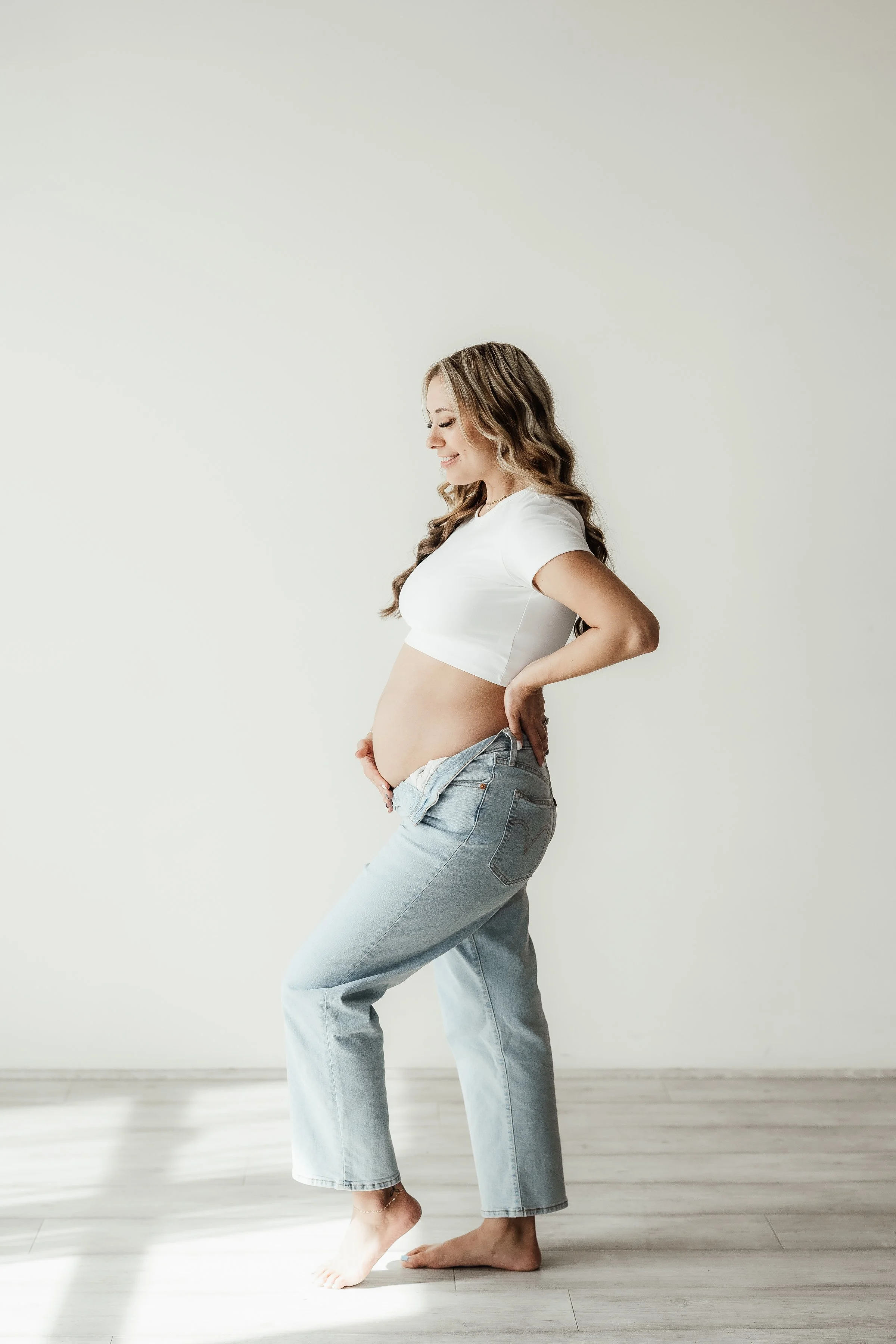 A pregnant woman in a white crop top and light blue jeans standing barefoot indoors against a plain light-colored wall, smiling and gently cradling her belly.