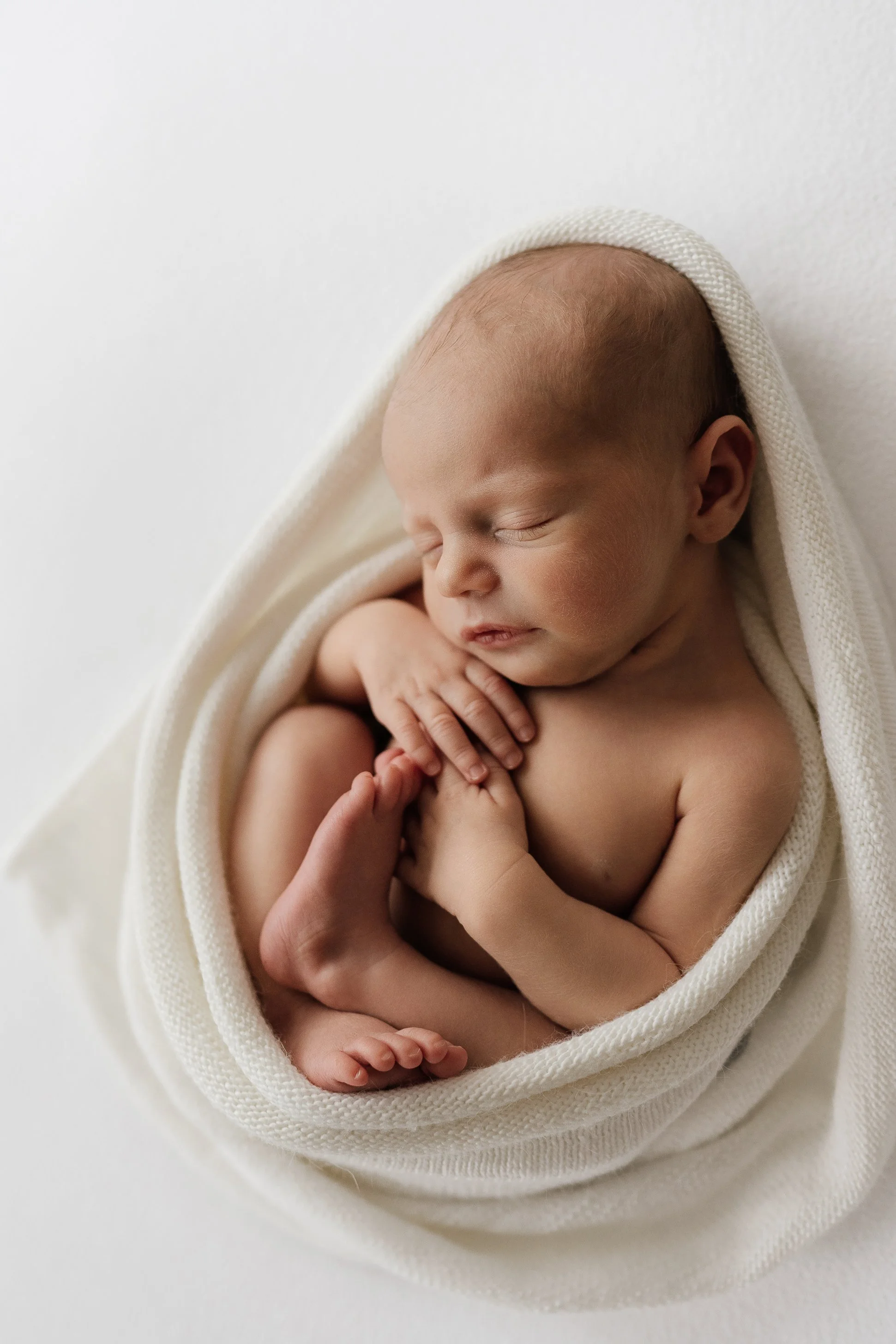 Newborn baby peacefully sleeping, wrapped in a cream-colored blanket, on a white background.