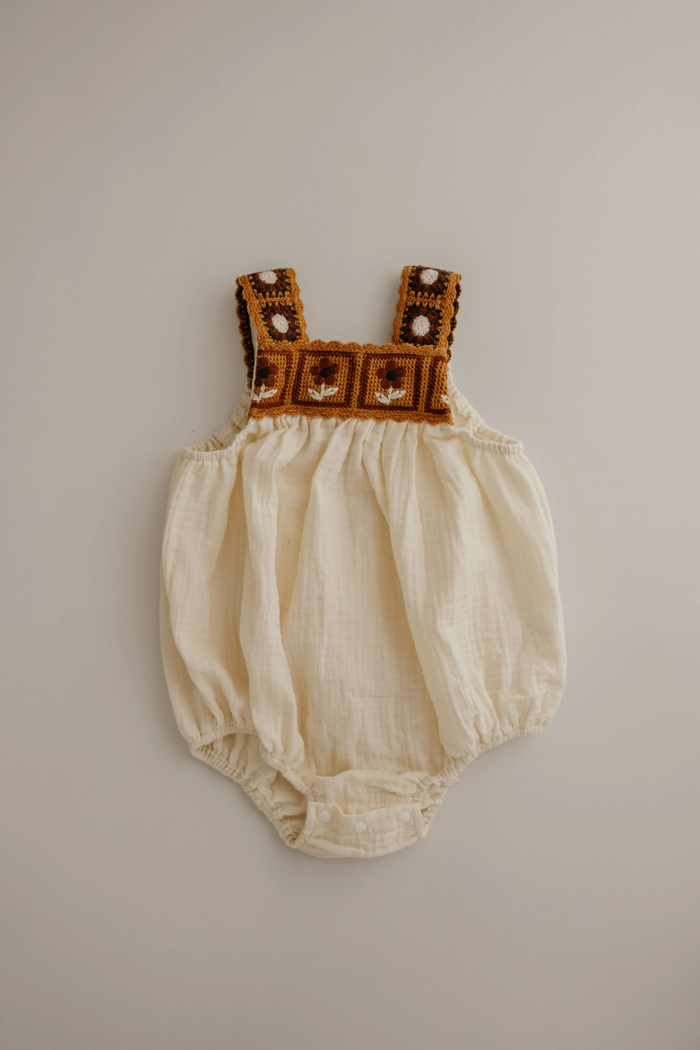 A vintage-style baby romper with a cream-colored fabric and decorative shoulder straps featuring floral embroidery.