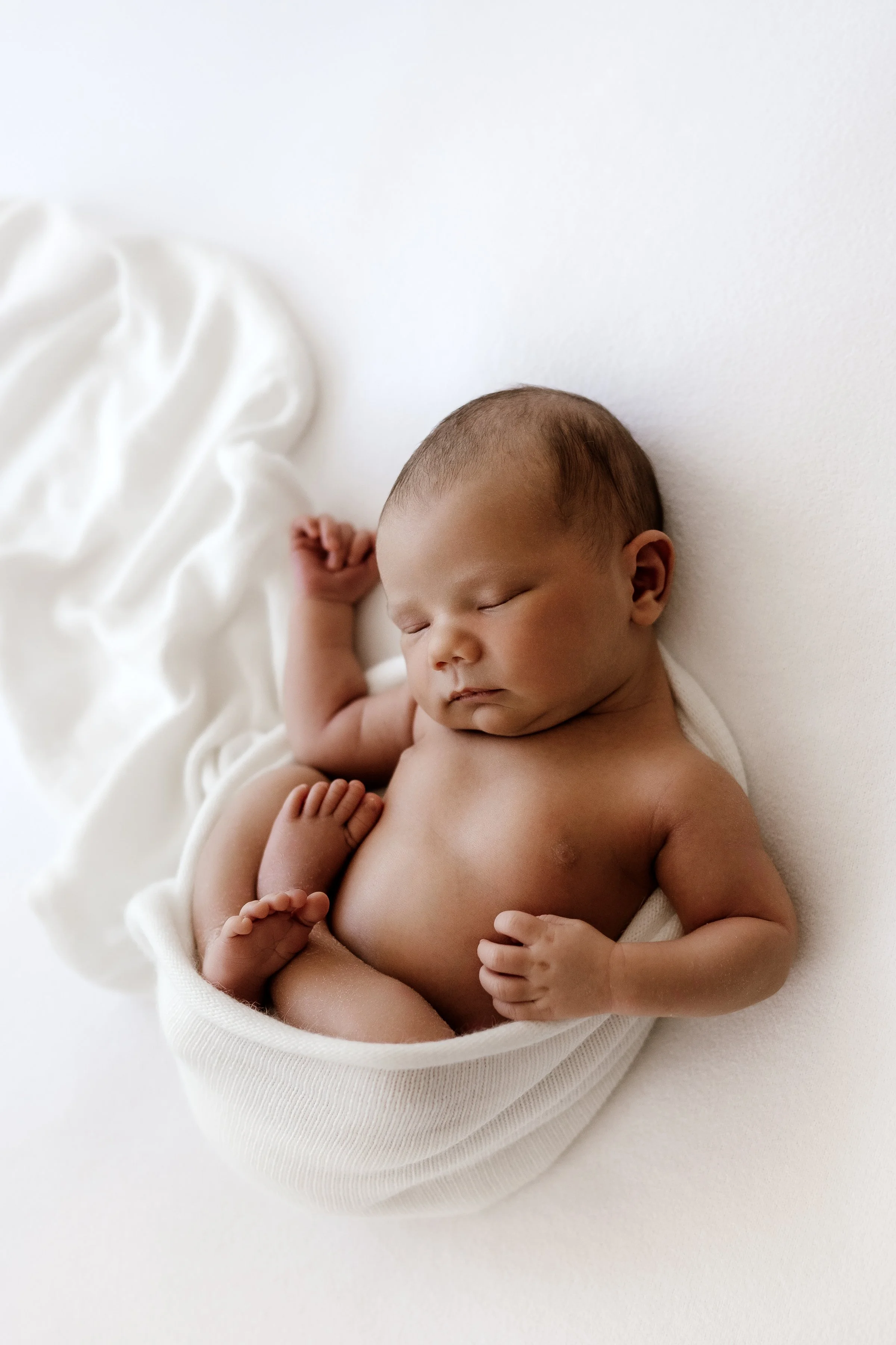 A sleeping newborn baby swaddled in a white cloth, lying on a white surface with a white blanket nearby.