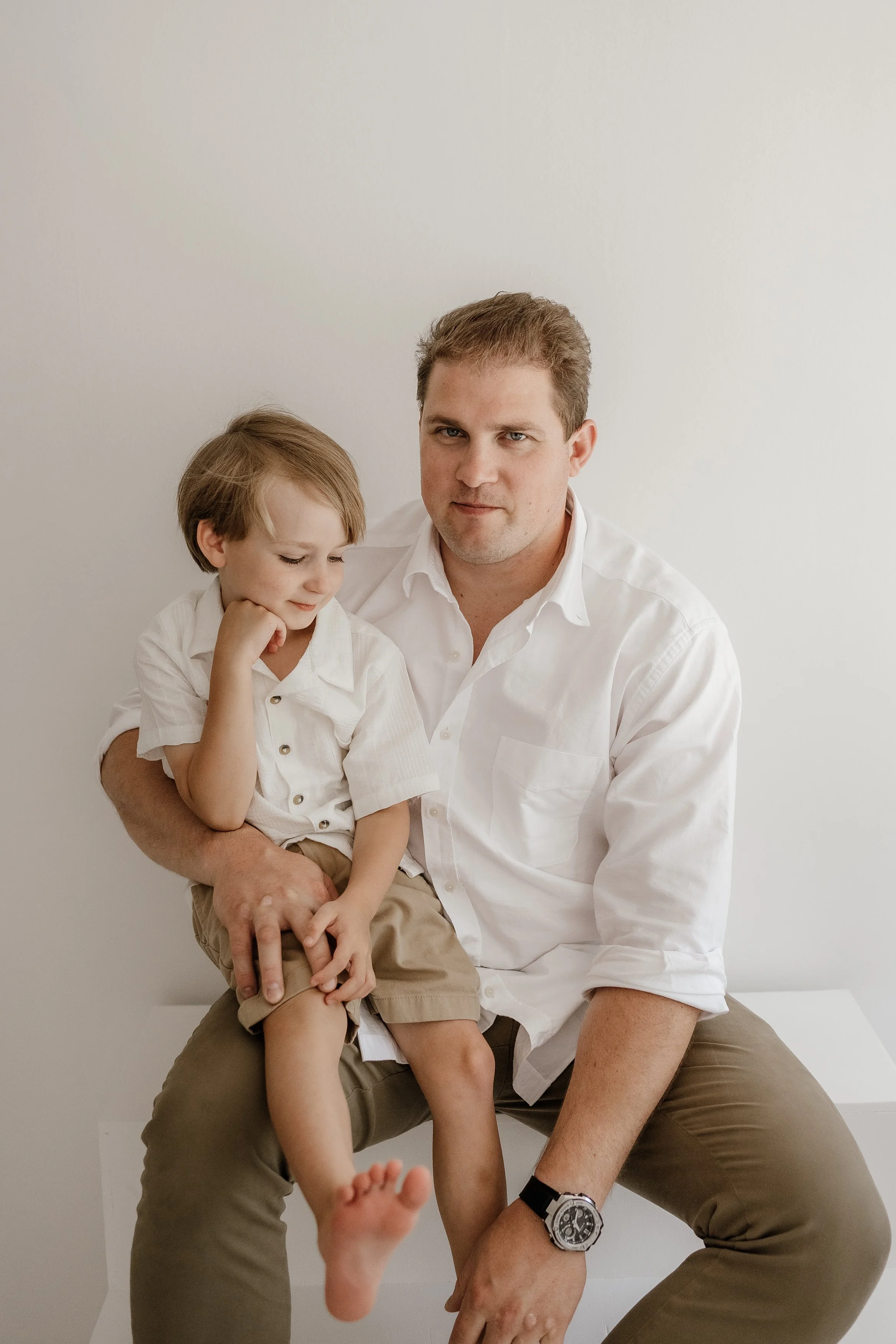 A man and young boy sitting against a plain white wall, with the man holding the boy on his lap. Both are wearing white shirts; the boy has a short-sleeved button-up and the man has the sleeves rolled up. The man has short, light brown hair and a ser