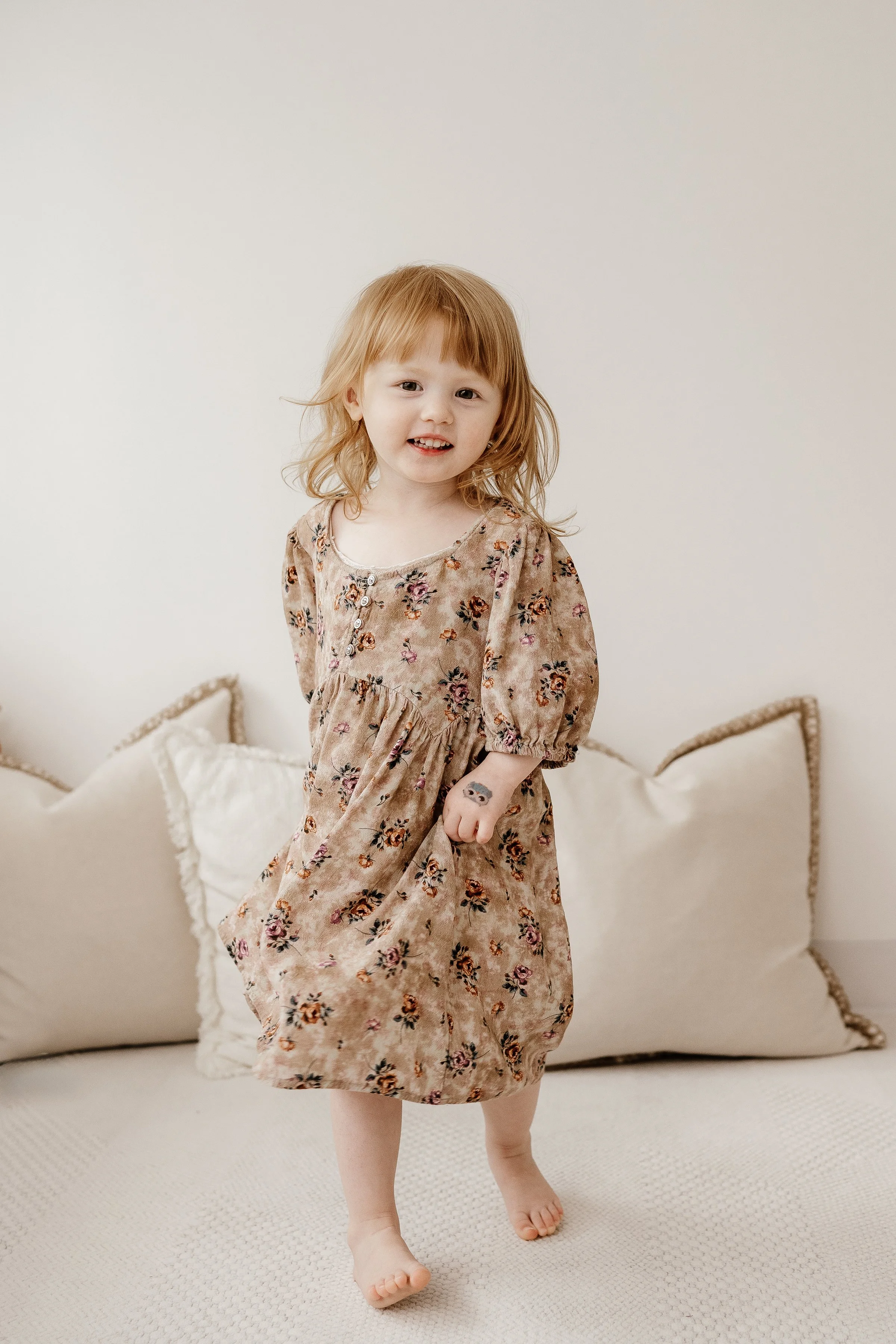 A young girl with red hair, wearing a floral dress, standing on a couch in front of a plain wall.