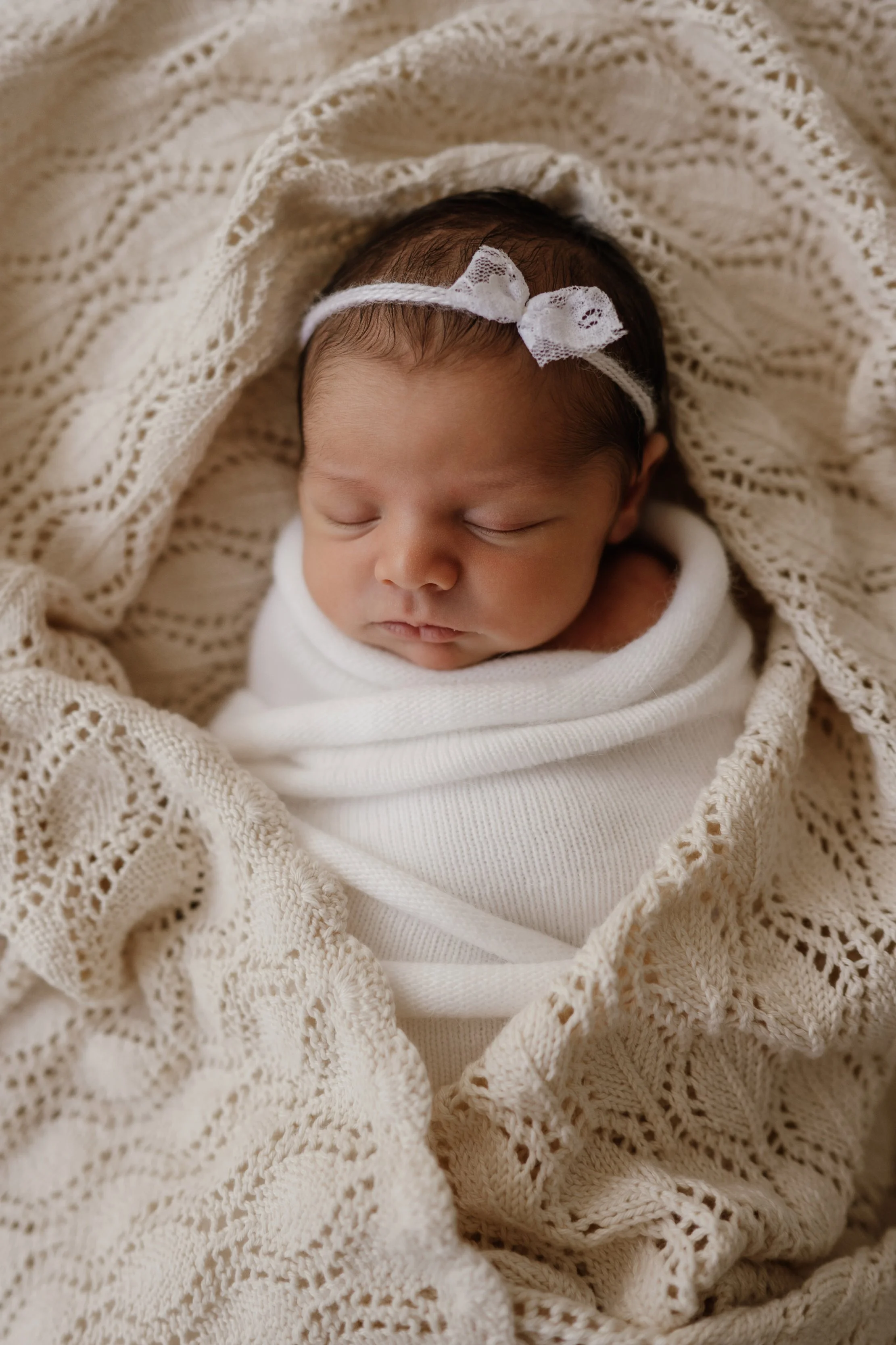 Close-up of a sleeping newborn baby wrapped in a cream-colored knitted blanket, wearing a white headband with a small bow.