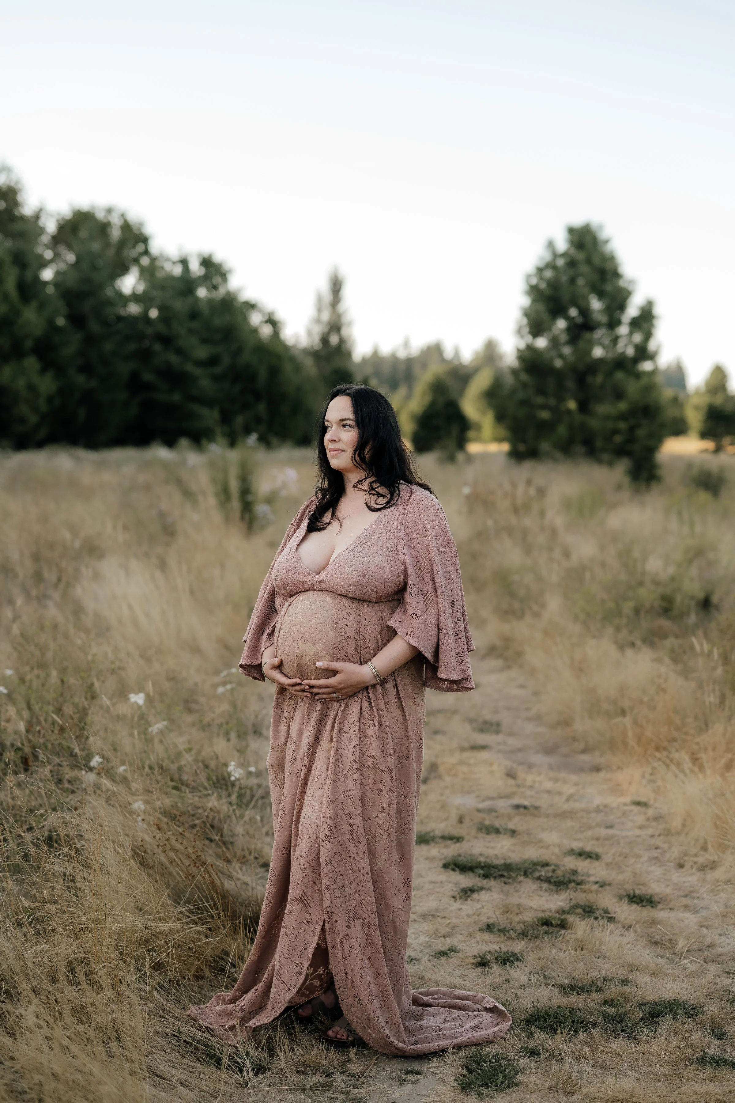 Pregnant woman in a pink lace dress standing on a dirt path in a grassy field with trees in the background.