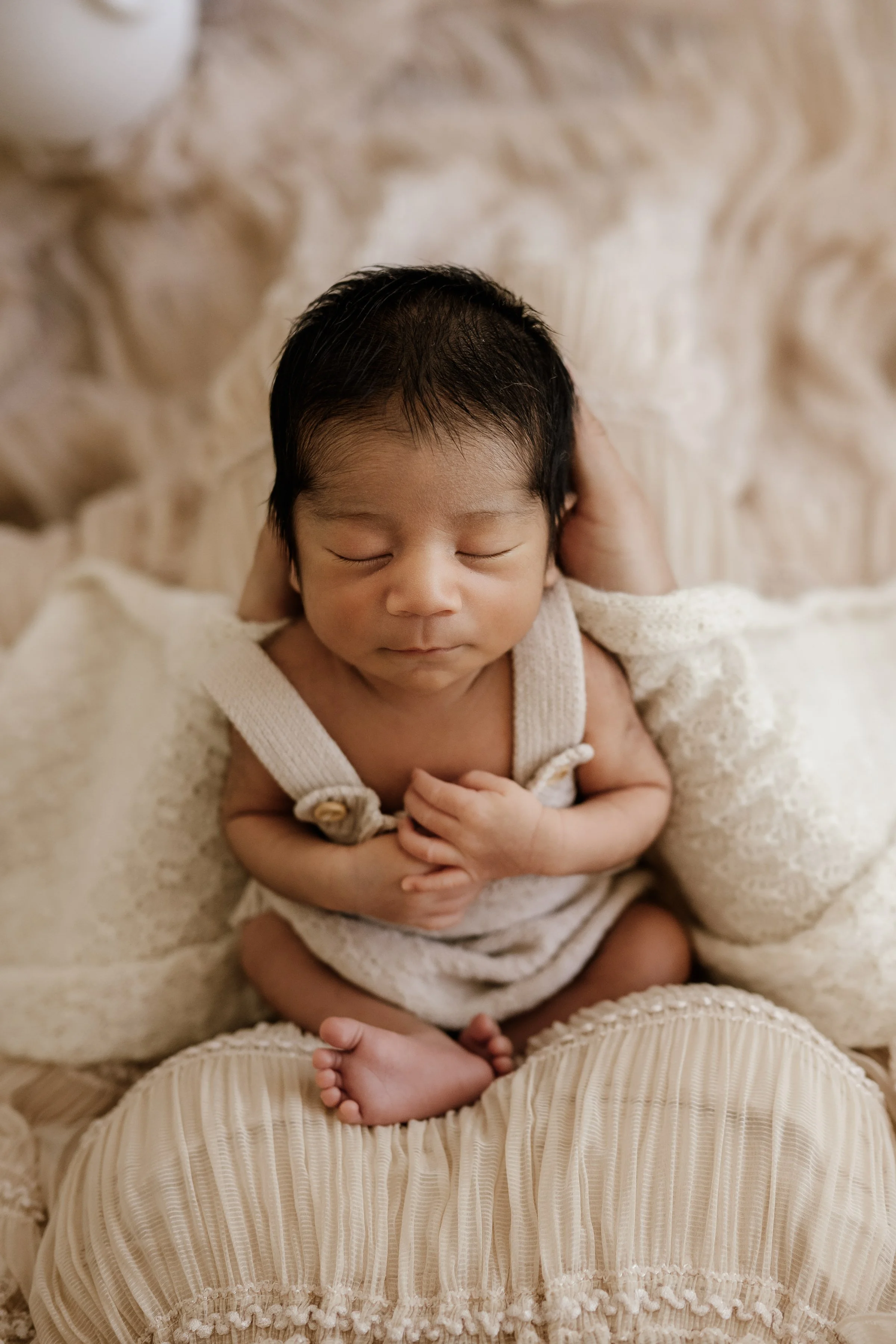 A newborn baby with dark hair sleeping peacefully, wrapped in cream-colored blankets, sitting on a person wearing cream-colored, textured, pleated pants.