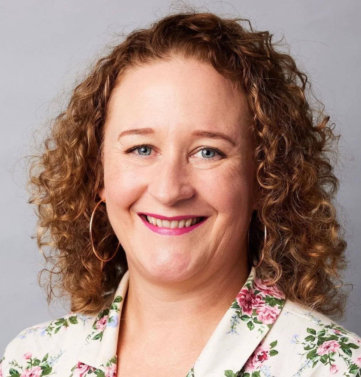 A photo of Erica (ADHD coach) with curly medium brown/red hair, blue eyes, and hoop earrings, smiling, wearing a floral blouse, against a plain grey background.