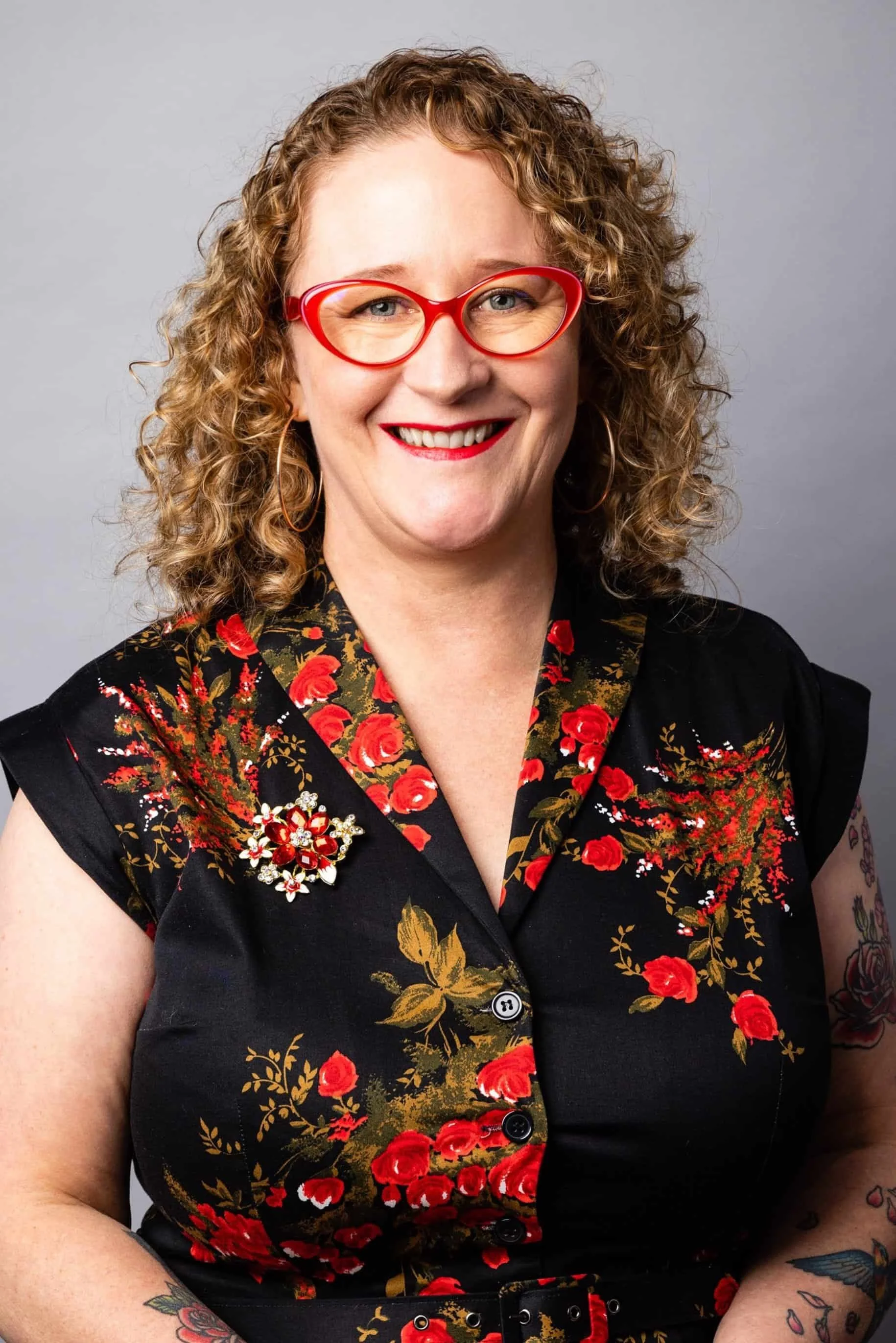 A photo of Erica, who offers ADHD coaching for women academics and professionals. She has shoulder length curly hair. She is smiling and has red glasses and lipstick and is wearing a shirt dress with red flowers with gold leaves.