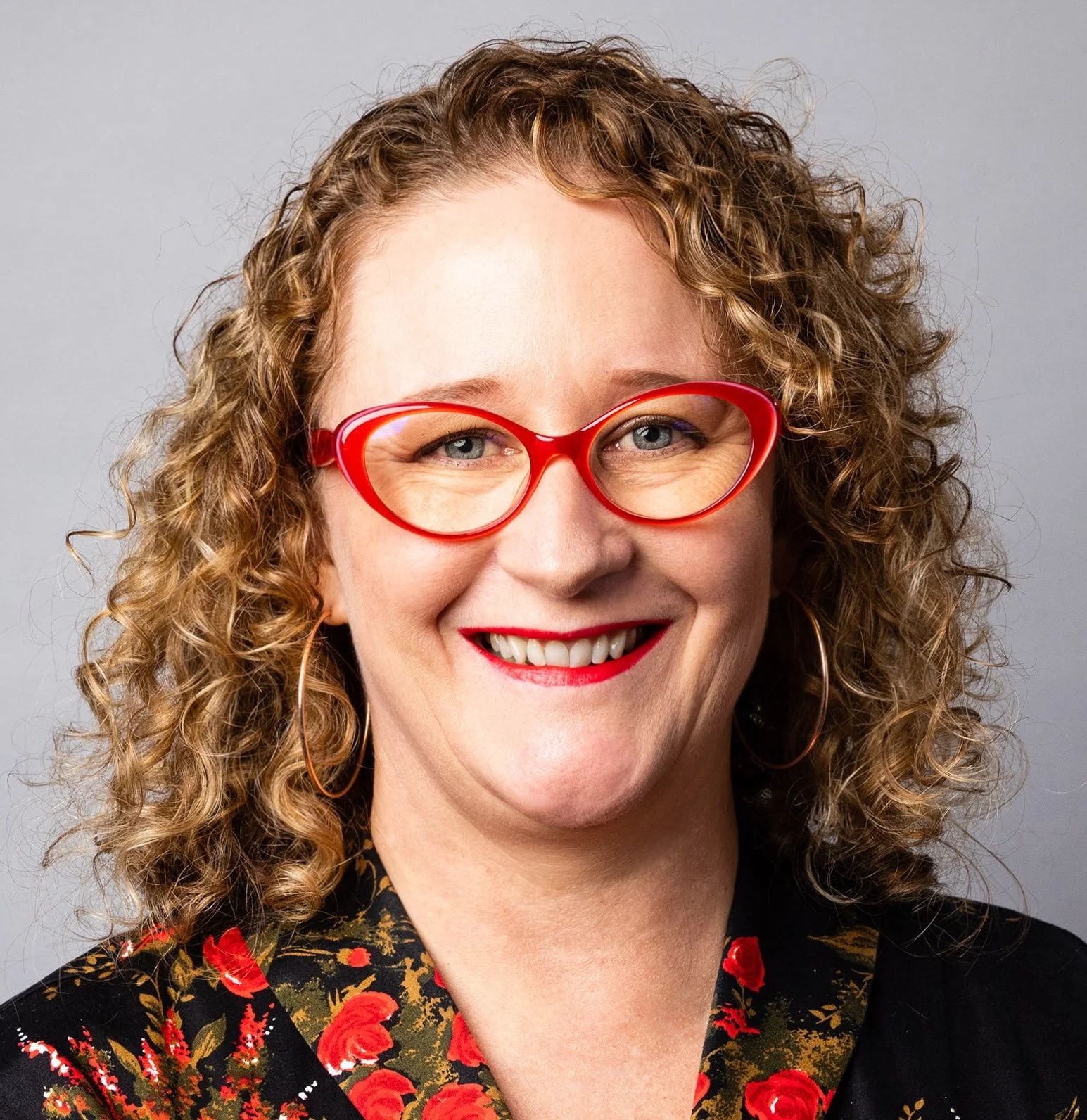 A photo of Erica with curly medium brown/red hair wearing red glasses, red lipstick, a black floral dress with red and gold accents, and a decorative brooch. She has tattoos on her arms and is smiling against a plain grey background.