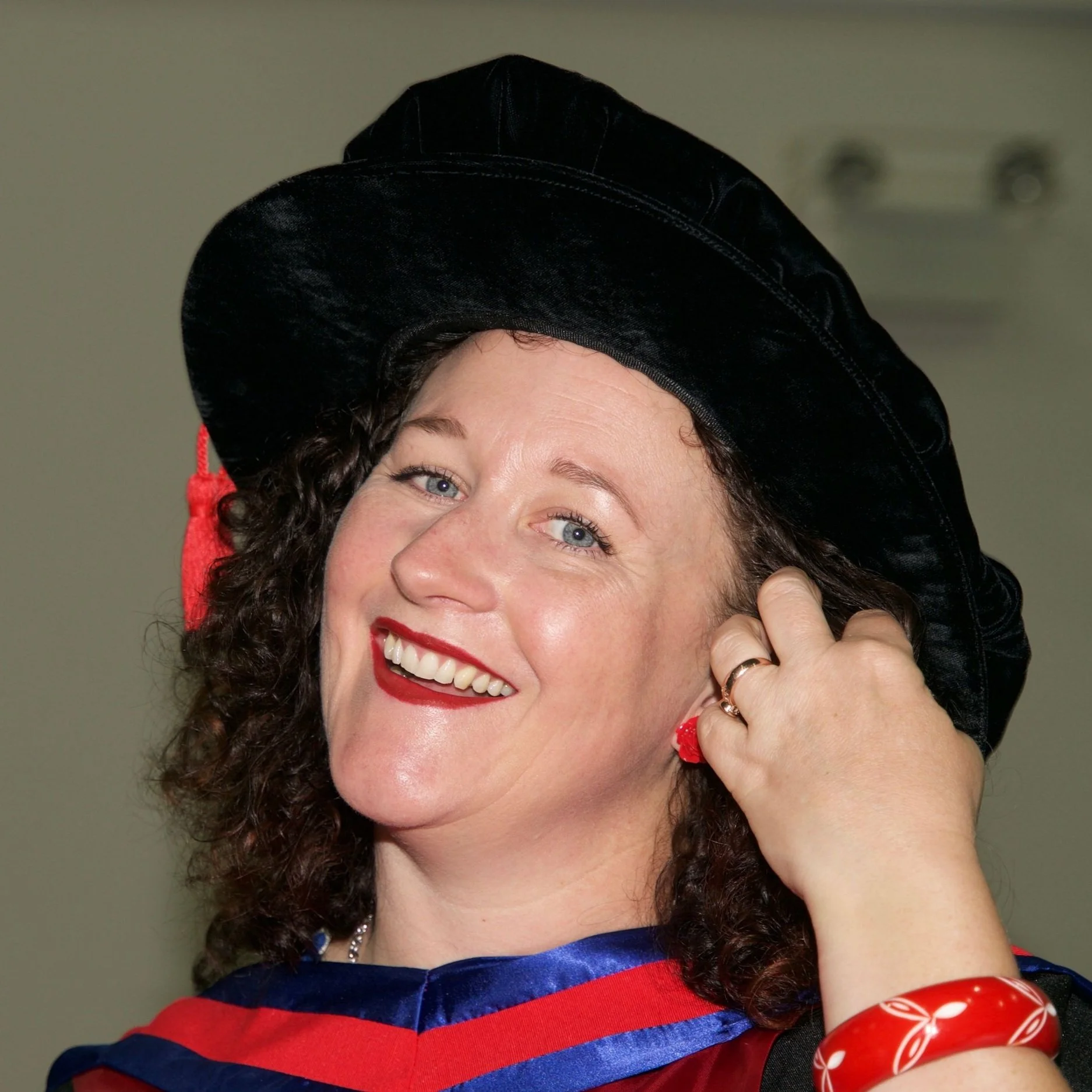 A photo of Erica with dark curly hair and red lipstick. She is smiling, wearing a graduation cap and gown, adjusting her hair.