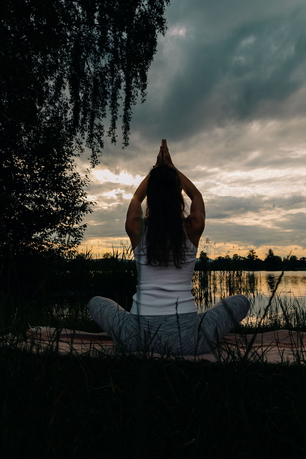 Frau, die im Lotussitz am Ufer eines Sees bei Sonnenuntergang, Pilates macht, mit den Händen über dem Kopf zusammengestellt.