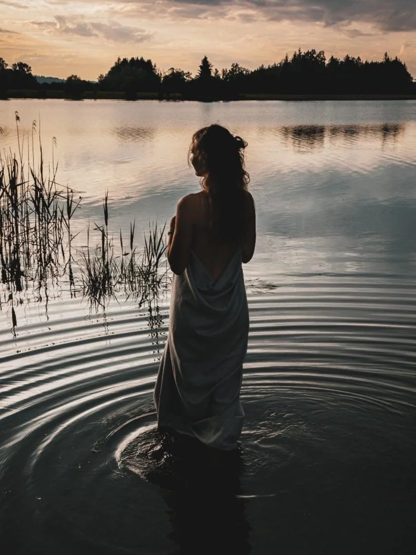 Eine Frau steht im Wasser an einem See bei Sonnenuntergang, umgeben von Natur und Bäumen im Hintergrund.