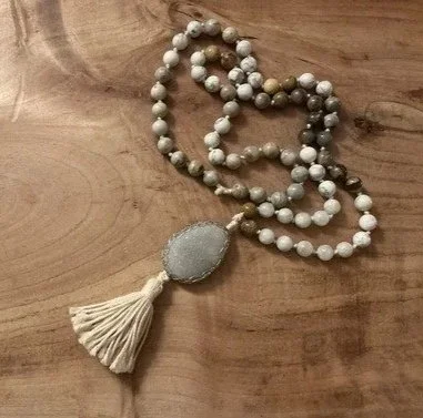 Eine Kette aus Mondstein, Howolith und Opalith Perlen, mit grauem Stein mit einer Quaste, die als Schmuckstück an einem Holztisch liegt. Boho-Kette, Boho-Schmuck