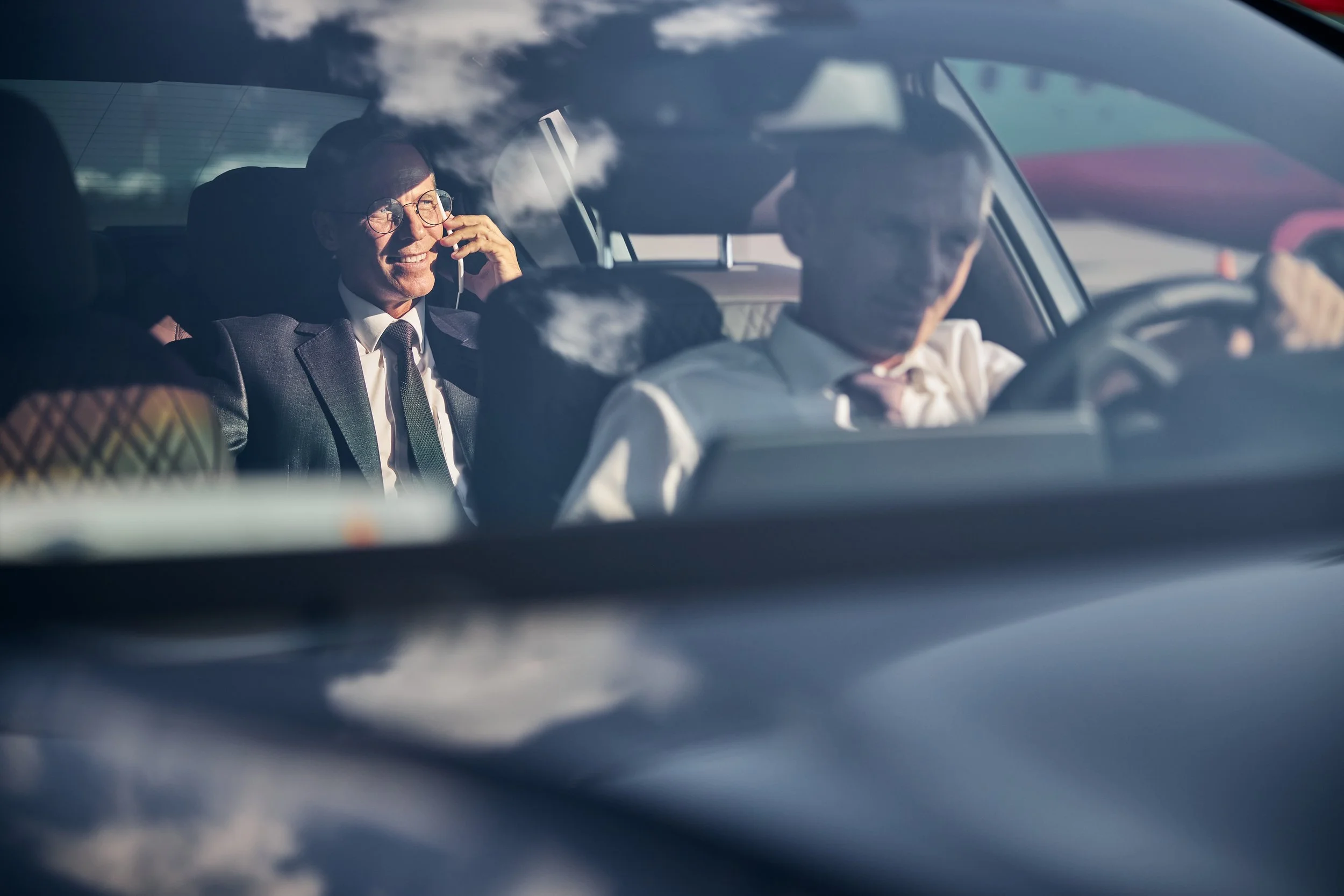 People inside a car, with one person smiling and talking on the phone, seen through a windshield with reflections of clouds.