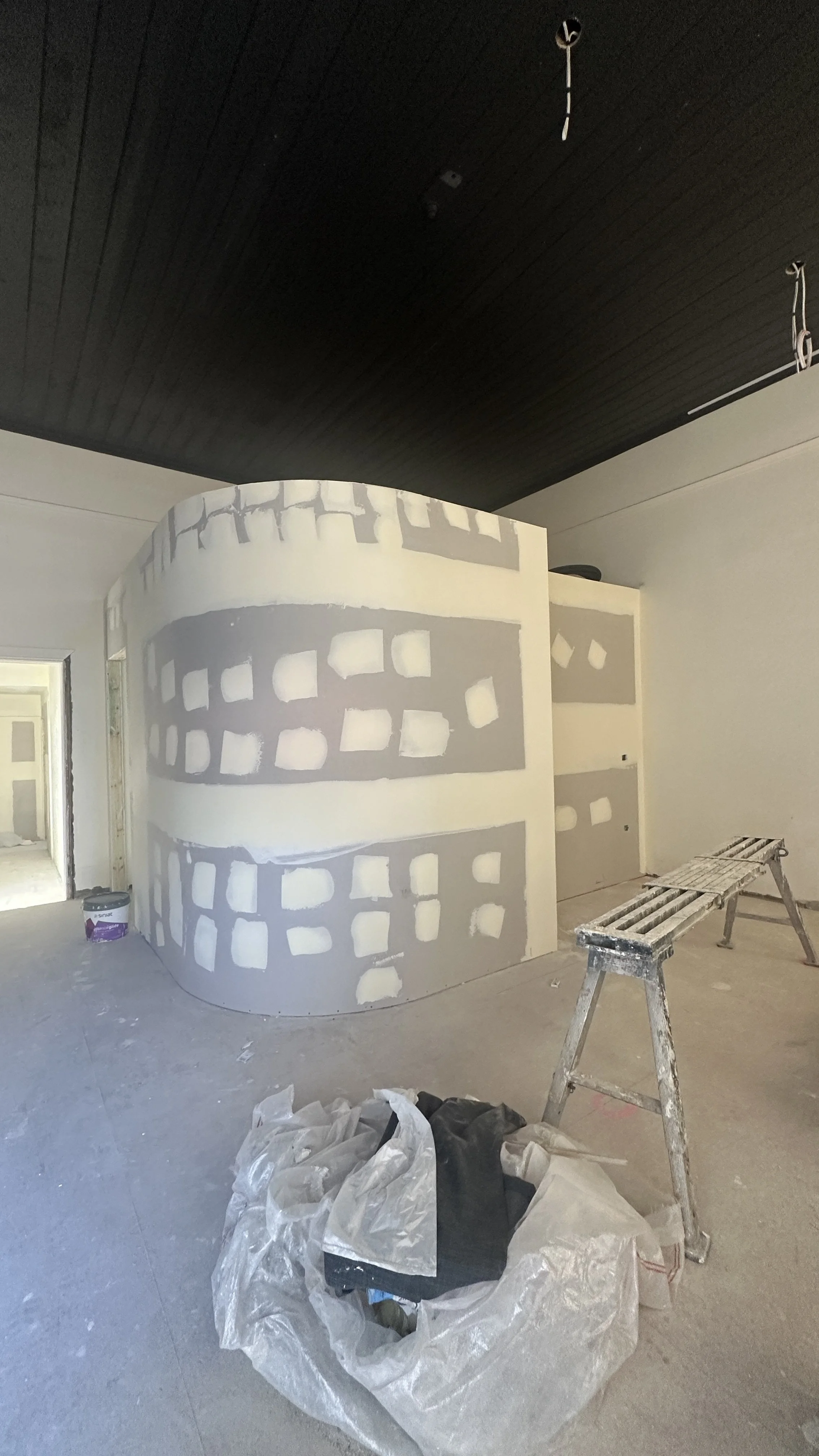 Modern medical office reception area with a marble counter, computer stations, and comfortable seating. Two women and a child wait near a large window, while a man reads papers. There is a digital signboard showing appointment booking options, plant decor, and warm lighting.