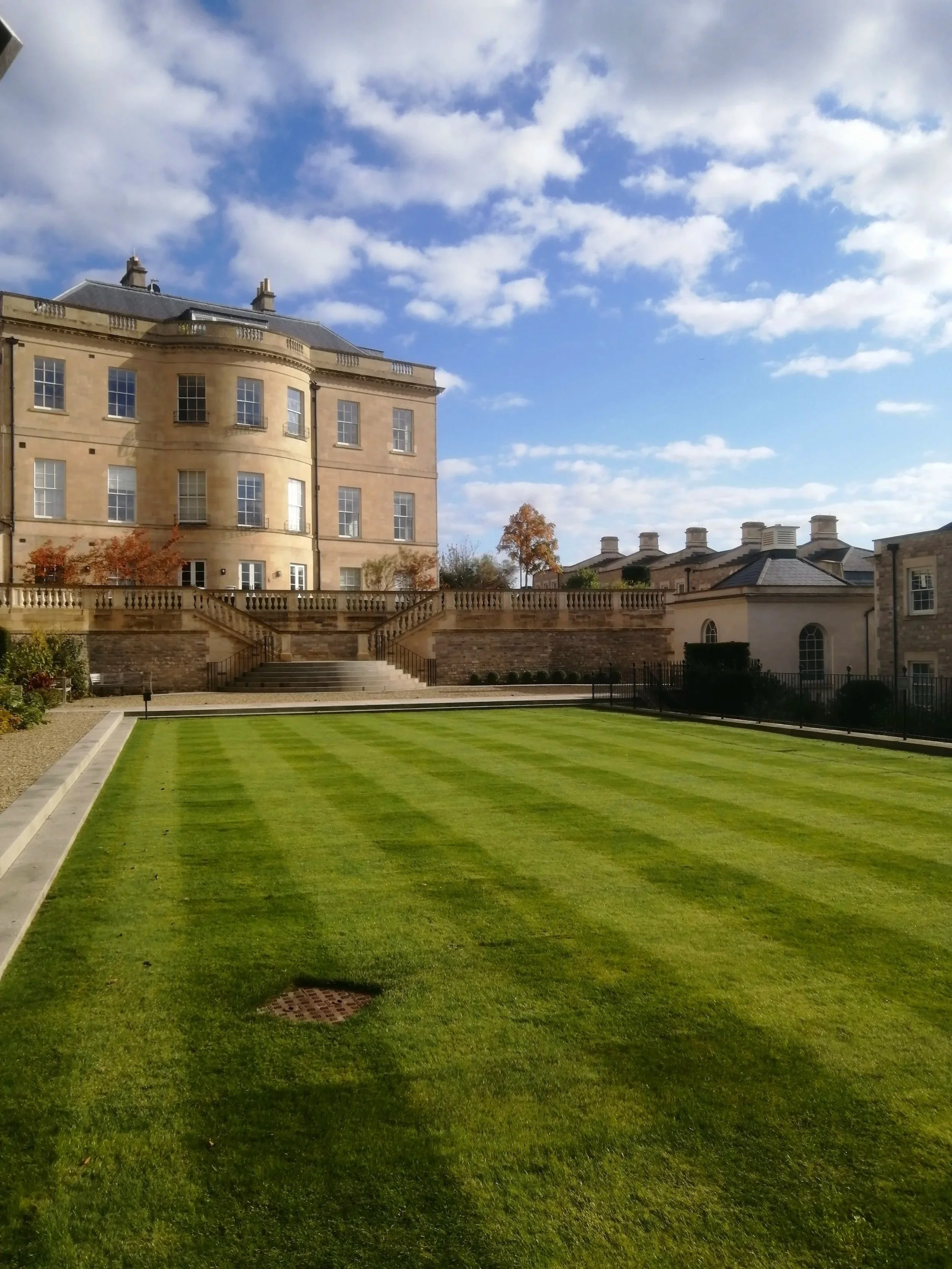 A well-maintained green lawn with a small drainage grate, in front of elegant stone buildings with multiple windows and a staircase. The sky is mostly cloudy with patches of blue.