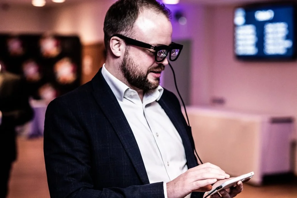 a photo of a man in a suit wearing smart glasses looking down at a whie mobile phone