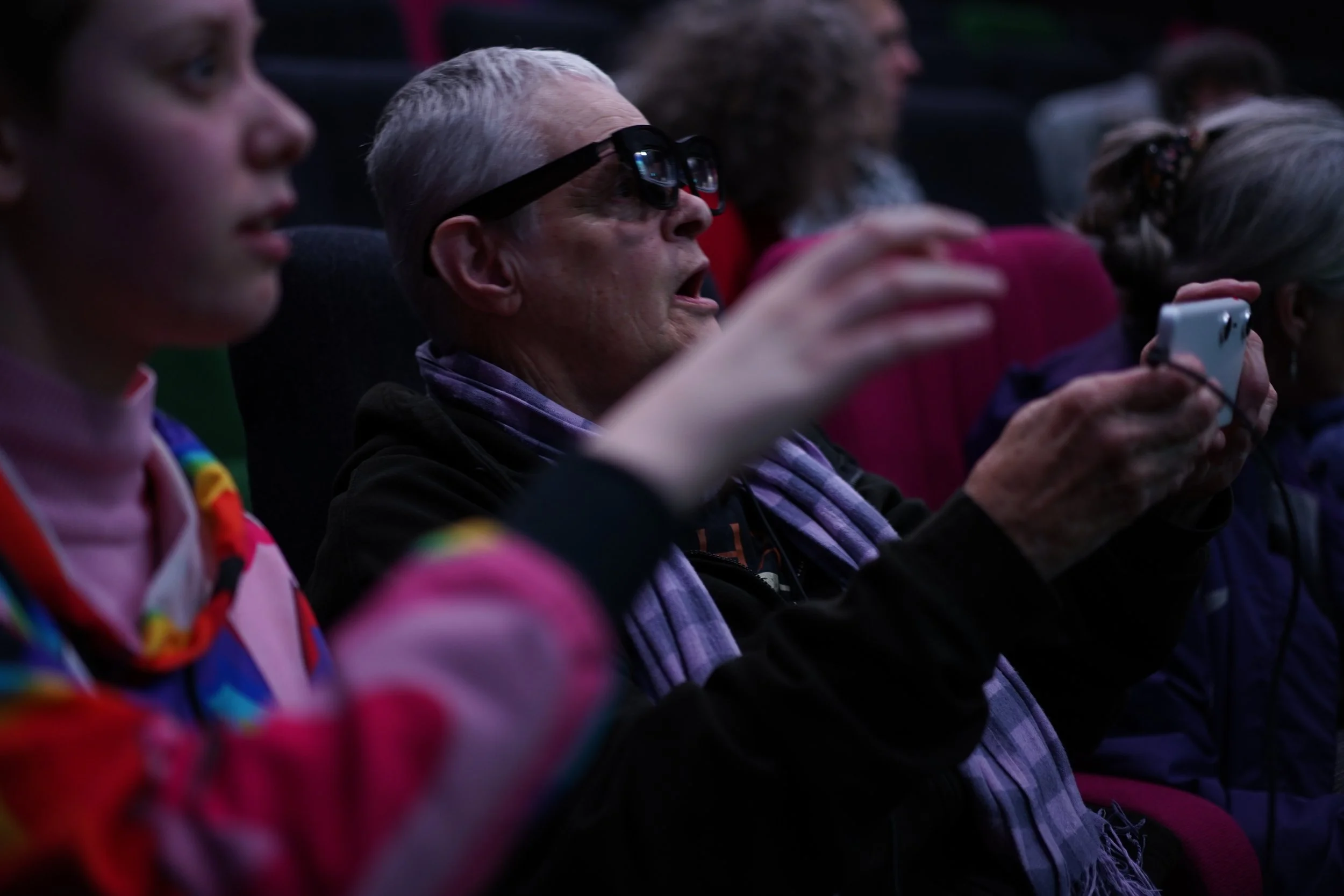 A close up of a woman with grey hair wearing a pair of smart glasses and holding a wired mobile phone up in front of her