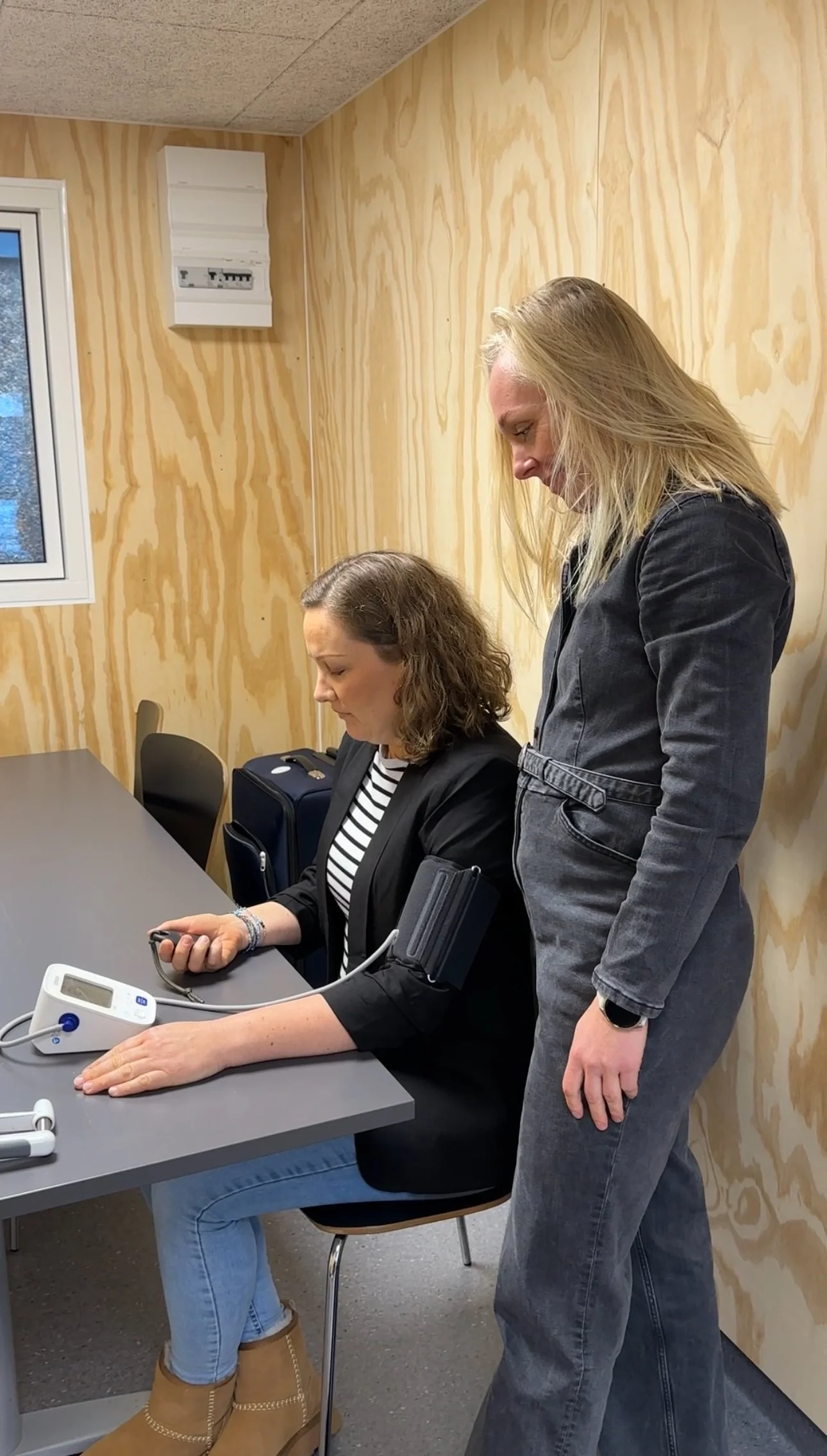 En kvinde sidder ved et bord med en blodtryksmåler og ser på sin telefon, mens en anden kvinde står ved siden af hende og overvåger blodtryksmålingen.