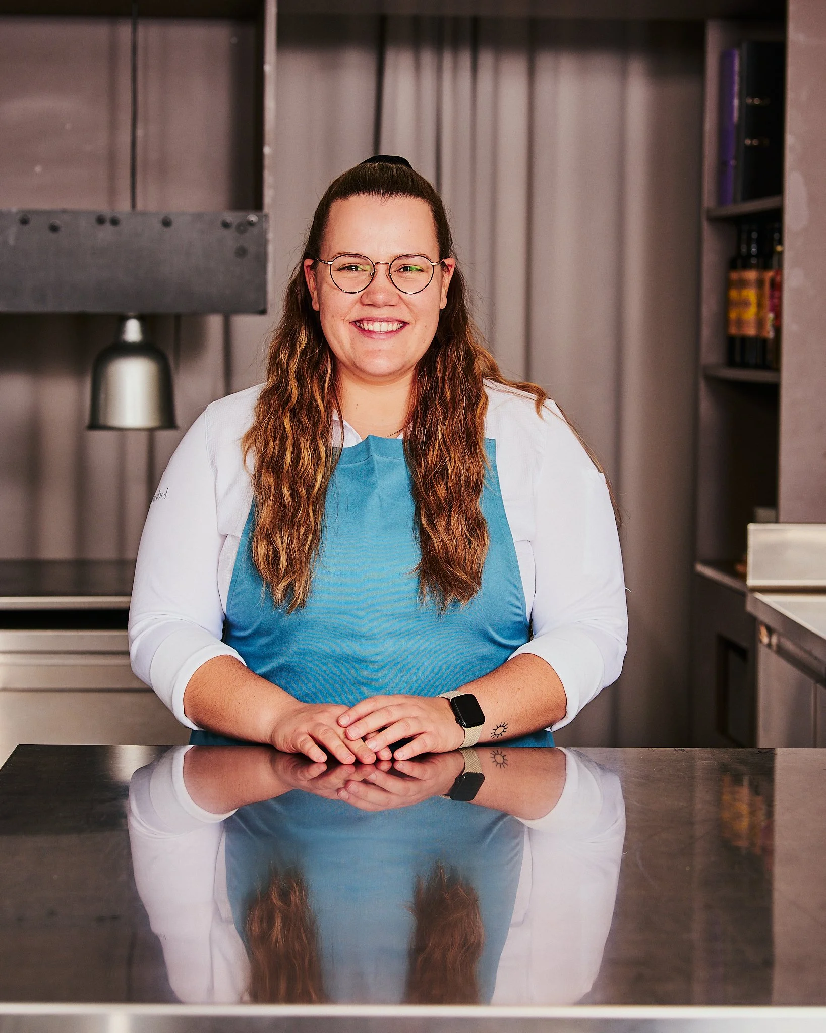 Frau mit Brille, braune Haare, blauer Kittel, steht im Küchenbereich mit metallischer Oberfläche und regt sich lächelnd