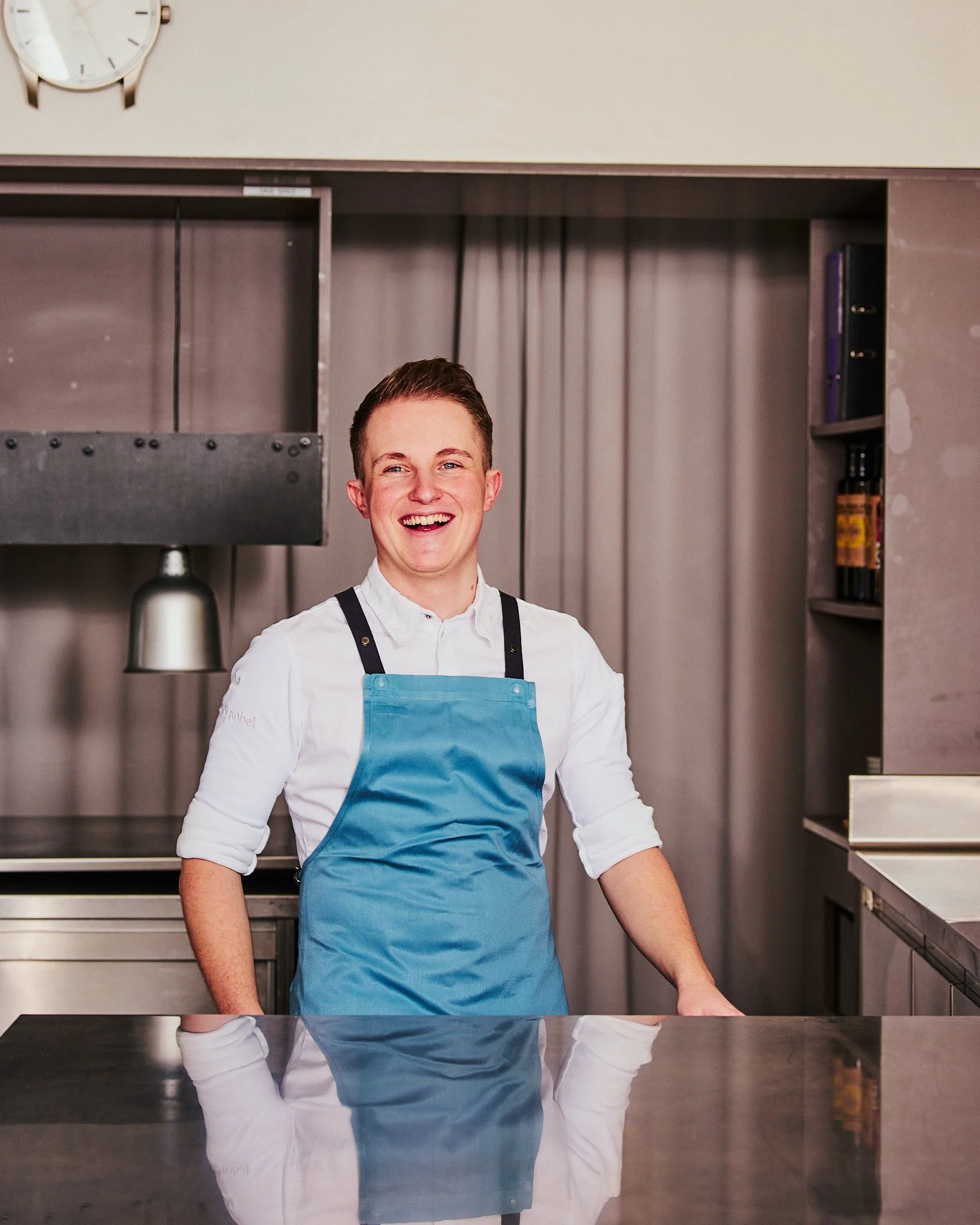 Ein junger Mann in Weiß und blauer Schürze, lachend in einer Küche.