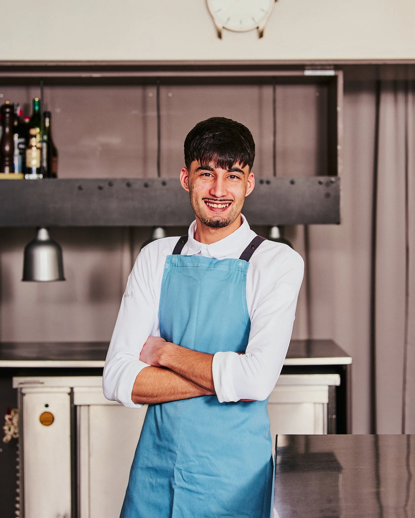 Ein Mann in Kochkleidung mit blauer Schürze steht mit verschränkten Armen in einer Küche und lächelt.