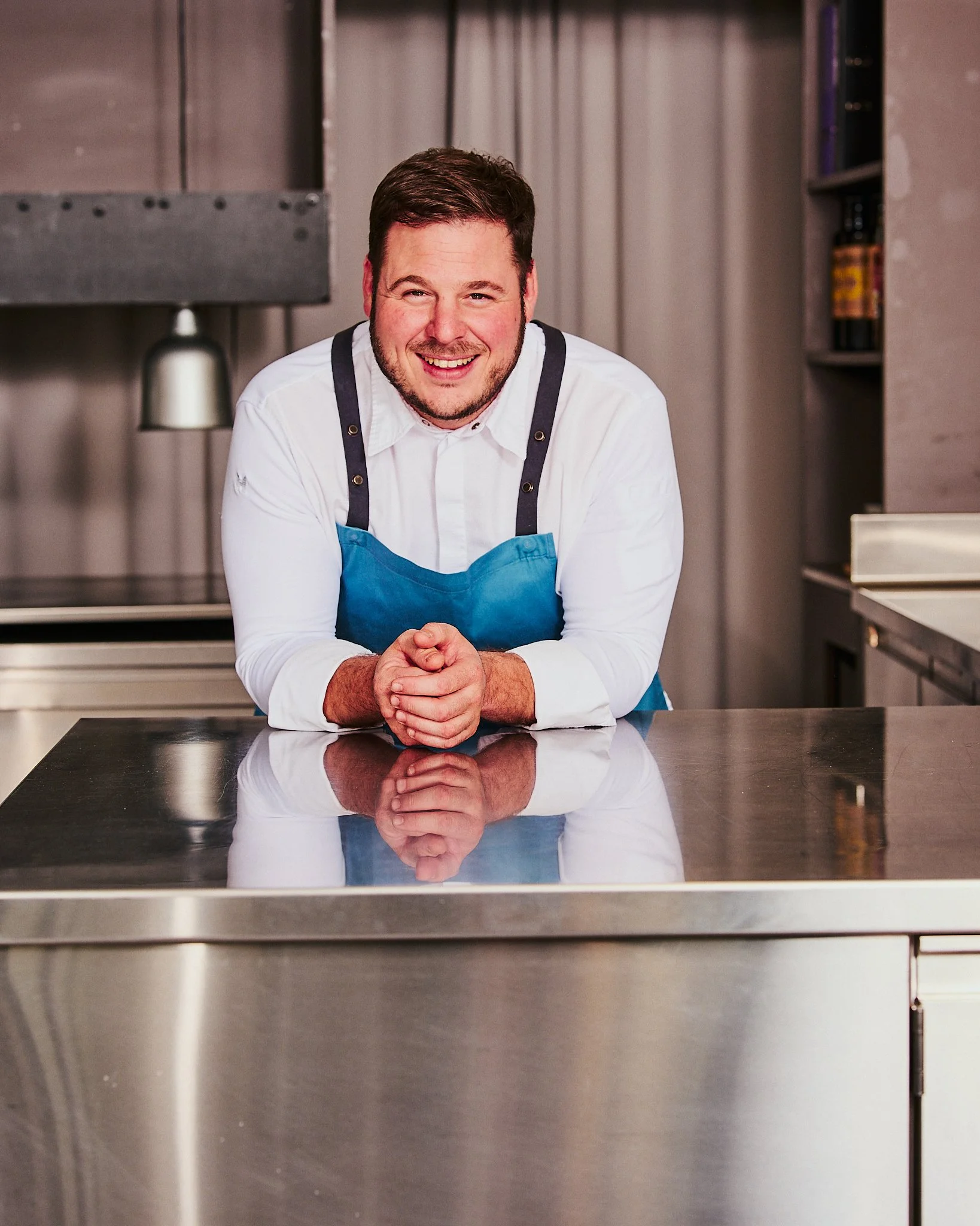Ein lachender Mann in weißem Hemd und blauer Kochschürze steht hinter einer Edelstahlküchentheke, die in einem professionellen Küchenumfeld ist.