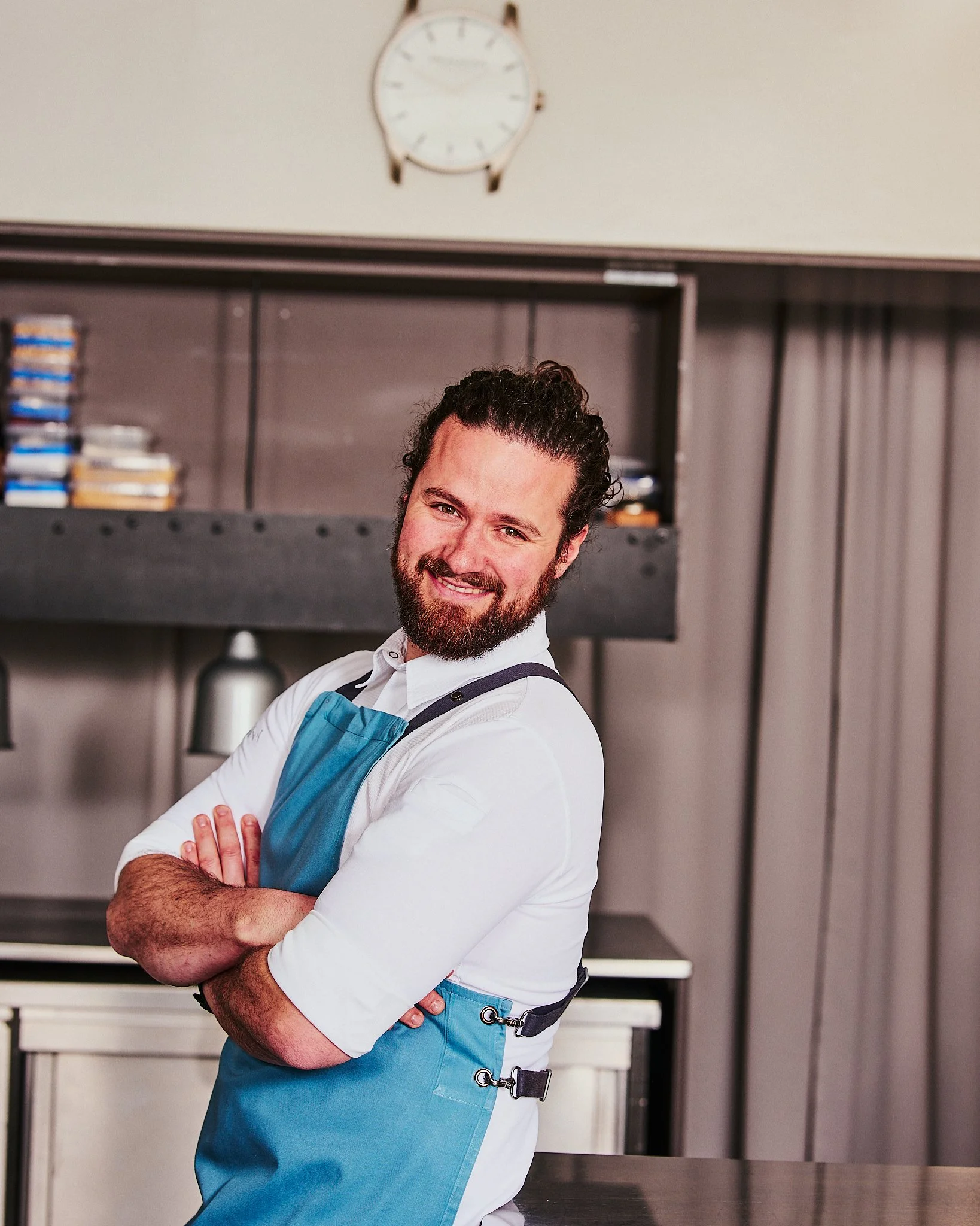 Ein fröhlicher Mann in einer blauen Kochschürze steht mit verschränkten Armen in einer Restaurantküche, im Hintergrund ein Regal und eine Uhr an der Wand.
