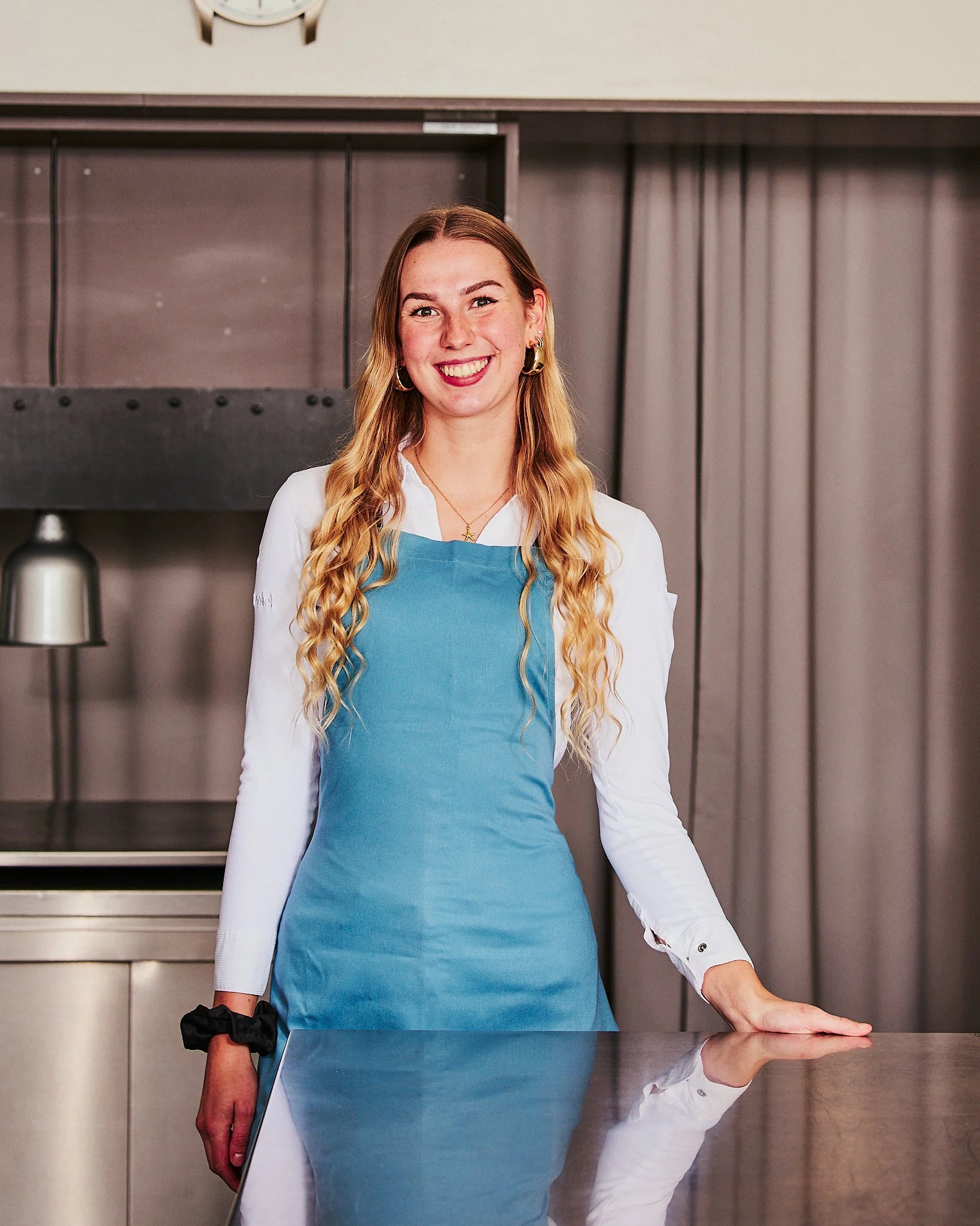 Eine lachende junge Frau mit lang, gewelltem, blondem Haar, trägt eine weiße Bluse und eine blaue Schürze, steht hinter einer Edelstahl-Arbeitsfläche in einer modernen Küche.