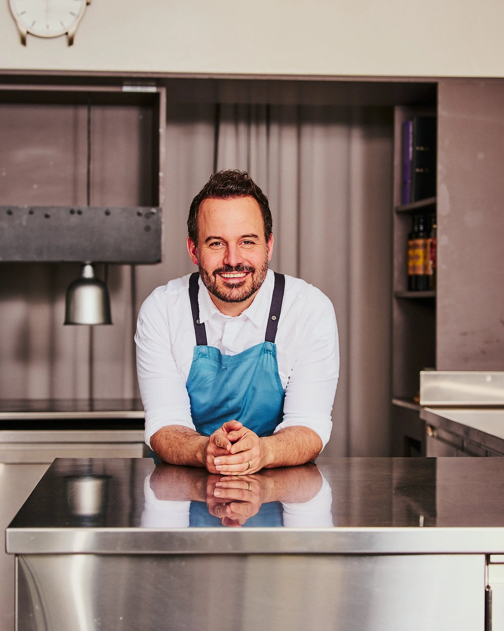 Ein Mann mit dunklem Haar und Bart, trägt eine weiße Hemd und eine blaue Schürze, steht hinter einer Metallarbeitsfläche in einer modernen Küche, lächelt freundlich ins Kamera.