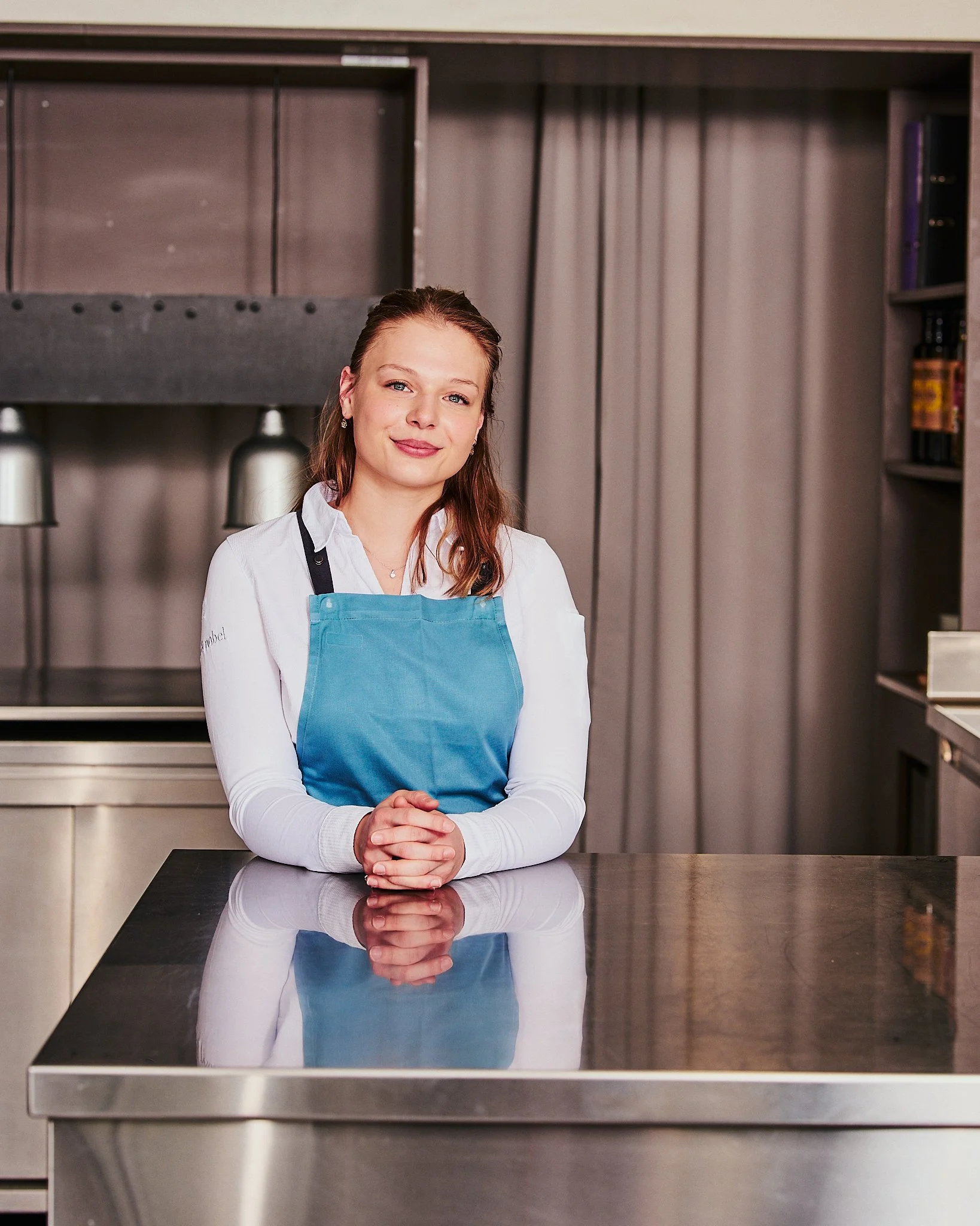 Eine junge Frau steht hinter einer Metalltheke in einer modernen Küche, trägt eine blaue Schürze und lächelt freundlich.
