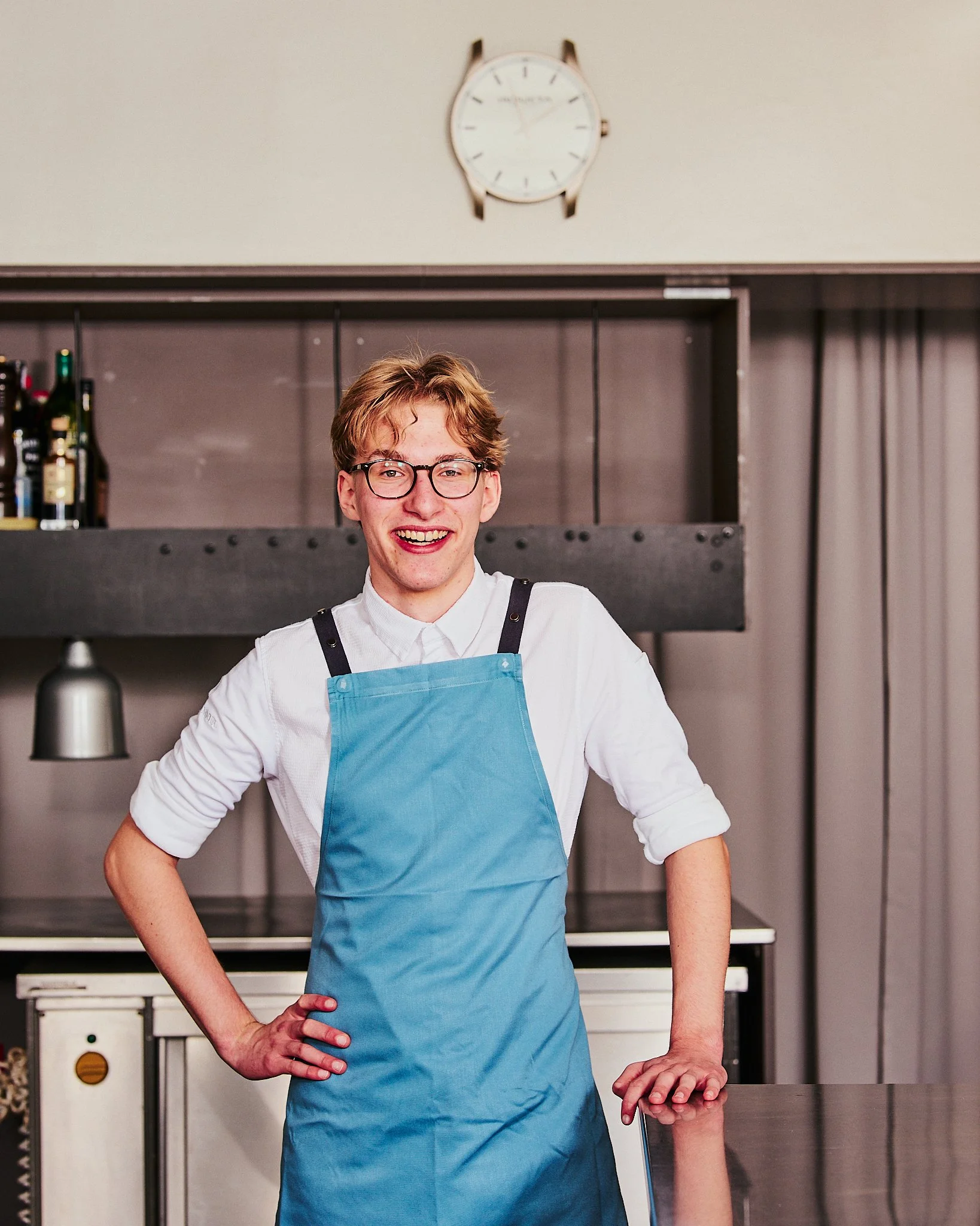 Ein fröhlicher junger Mann mit Brille, weißem Hemd und blauer Schürze steht in einer modernen Küche, lächelt in die Kamera.