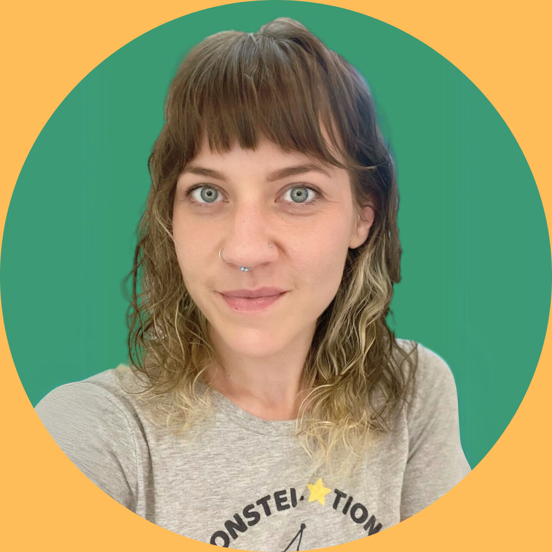 A woman with shoulder-length wavy hair, blue eyes, and nose and septum piercings, smiling at the camera, wearing a gray t-shirt with a partial logo on it, against a green background with a yellow border.
