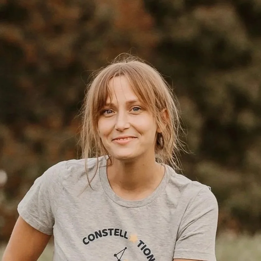 A woman with light brown, messy hair smiling outdoors with a blurred natural background.