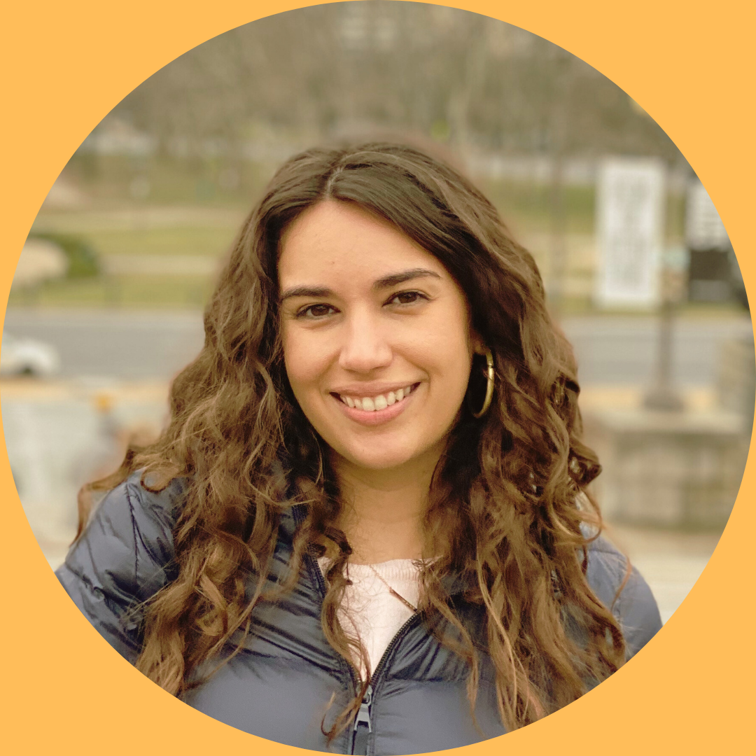 A smiling woman with long curly brown hair, wearing hoop earrings and a navy jacket, outdoors with a blurred background of a park and road.