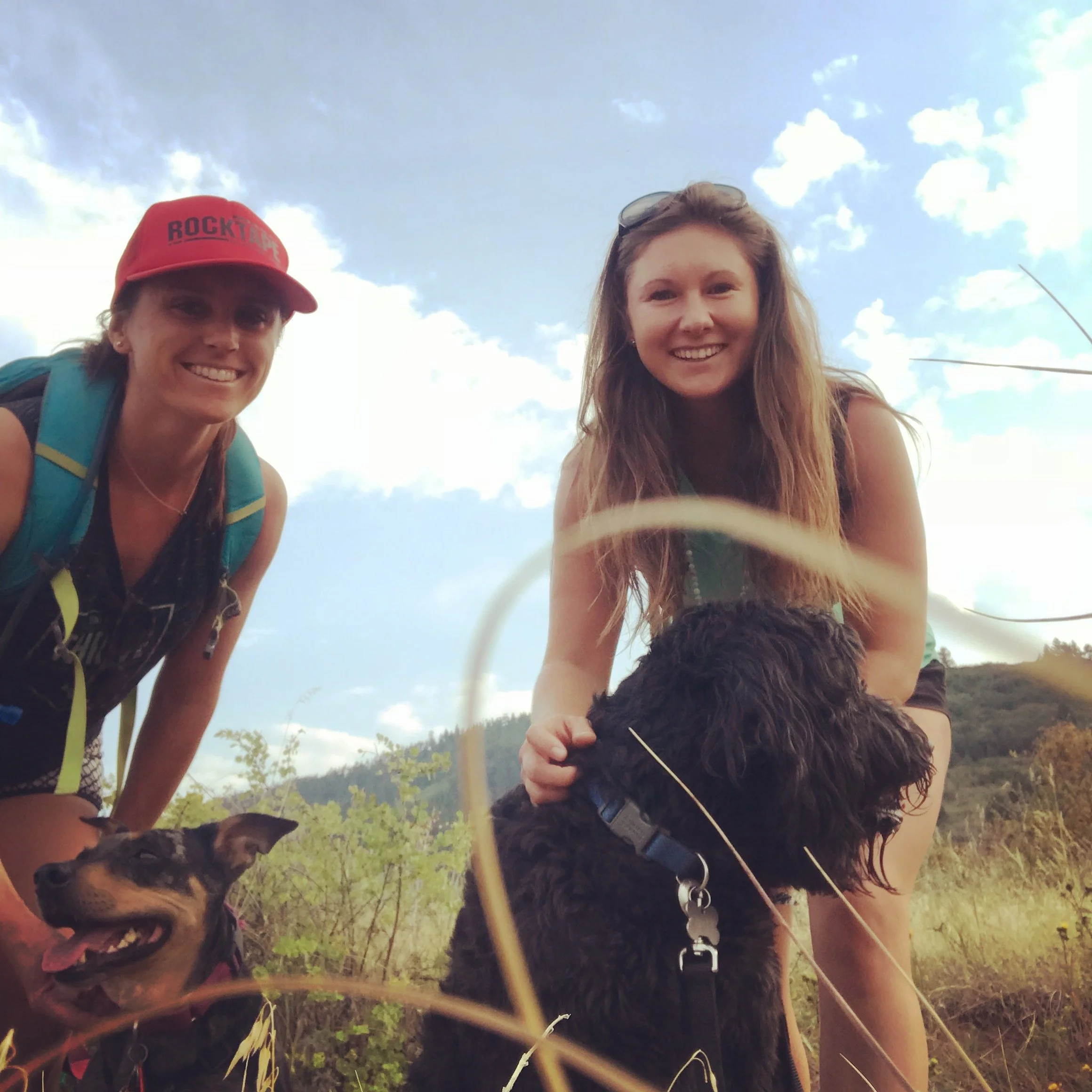 Two women and two dogs outdoors, smiling at the camera with a mountain and cloudy sky in the background; one woman is wearing a red cap with "ROCK" written on it, and both women are dressed casually.