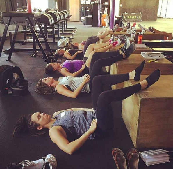 Group of women lying on their backs with legs elevated on wooden boxes during a workout class in a gym.