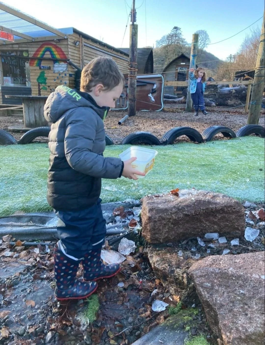 A young boy in a puffy coat and rain boots stands on wet ground near a large rock, holding a plastic container and looking down. In the background, a girl on a small rope bridge is playing in an outdoor play area with wooden structures, tires, and a mural of a rainbow on a building.