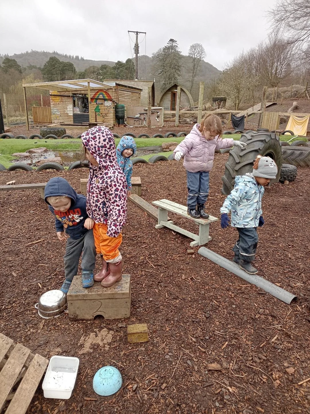 Children playing on outdoor play equipment made of wood and metal, dressed in rain jackets and hats, outdoors on a muddy ground with a farmyard background and cloudy sky.