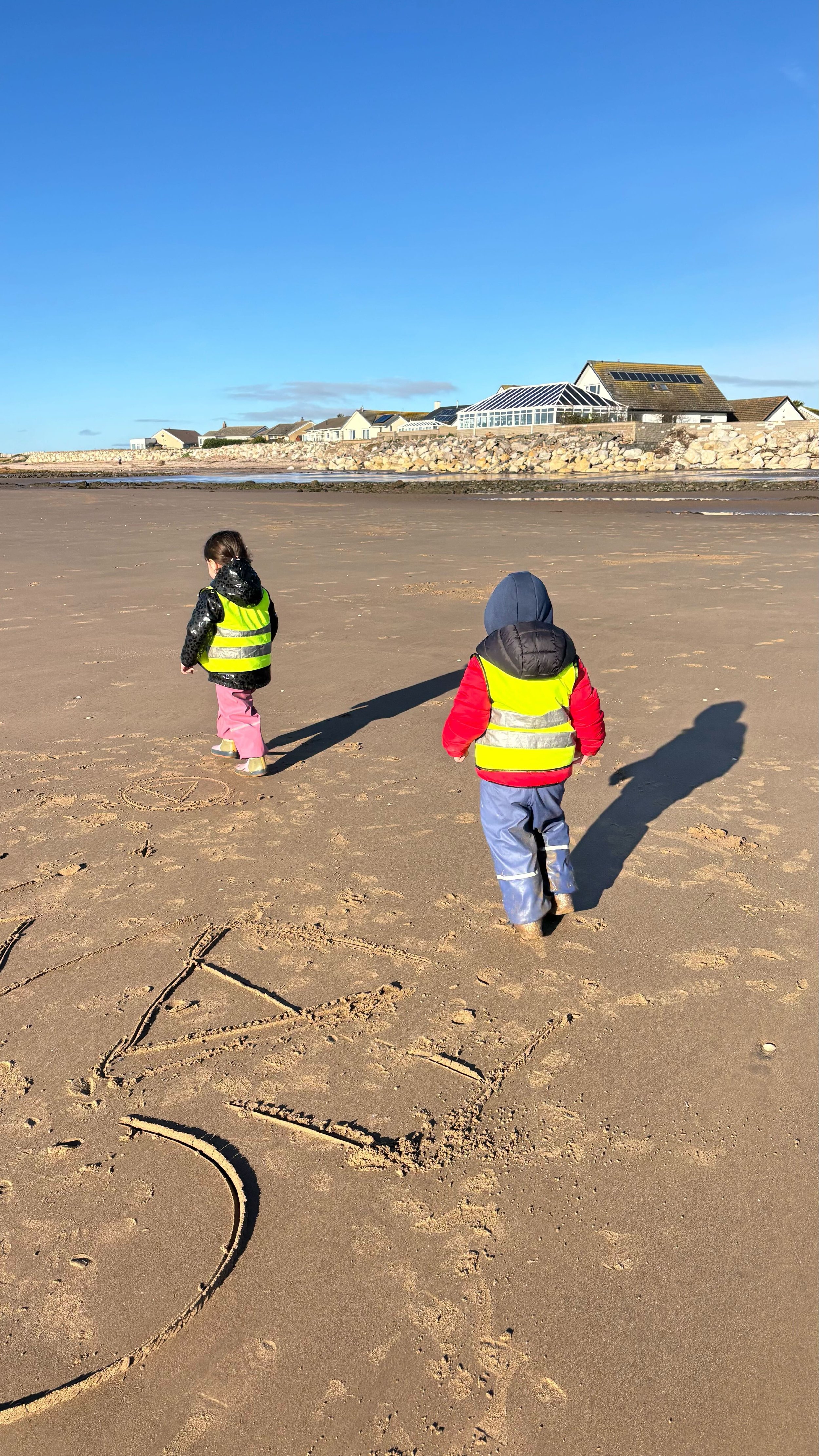 Two children walking on a sandy beach, wearing yellow safety vests. The boy is wearing a red jacket and blue pants with a hood, while the girl is in a black hoodie and pink pants. There are houses with solar panels on the roofs on a rocky embankment in the background, under a blue sky.