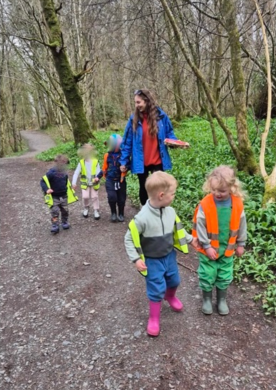 A woman and five children walking on a forest trail. The woman is holding some books and is talking to the children, who are wearing colorful rain boots and jackets, some with reflective vests.