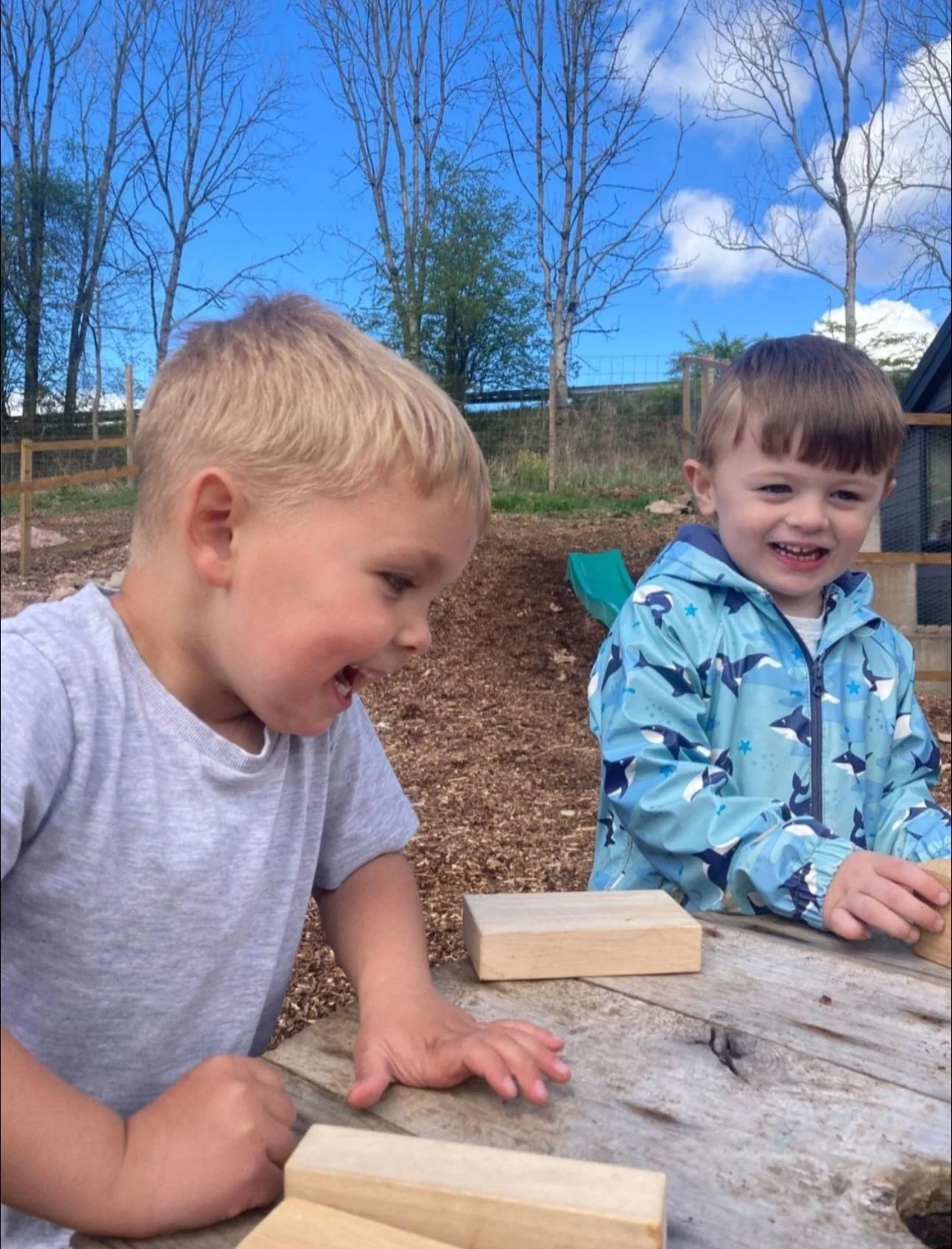 Two young boys playing outdoors at a wooden table on a dirt surface, with trees and a fence in the background, one boy smiling and the other looking down, under a partly cloudy sky.