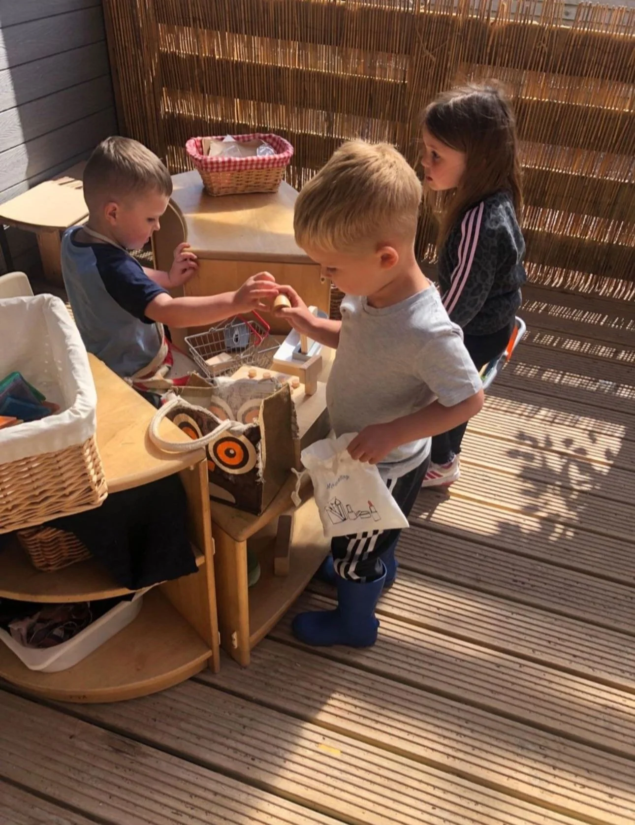 Four children playing with toys at a wooden table on a deck, with a bamboo privacy screen behind them. One boy is wearing blue rain boots and holding a bag, another boy is reaching into a small shopping cart, and a girl with dark hair stands observin