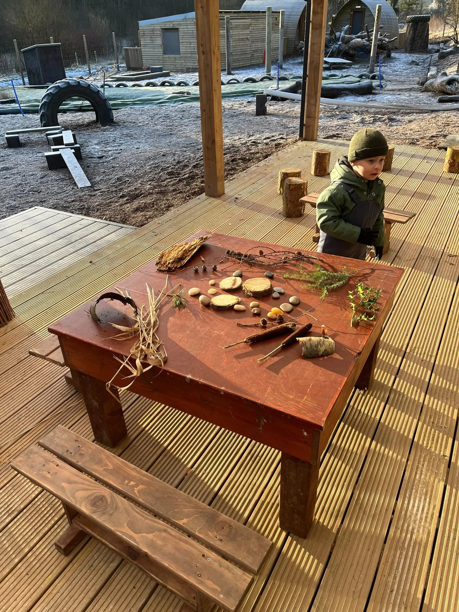 An outdoor playground with wooden and rubber obstacle course equipment. There are logs, tires, and a small boy in winter clothing standing near a table with natural objects like branches and stones.