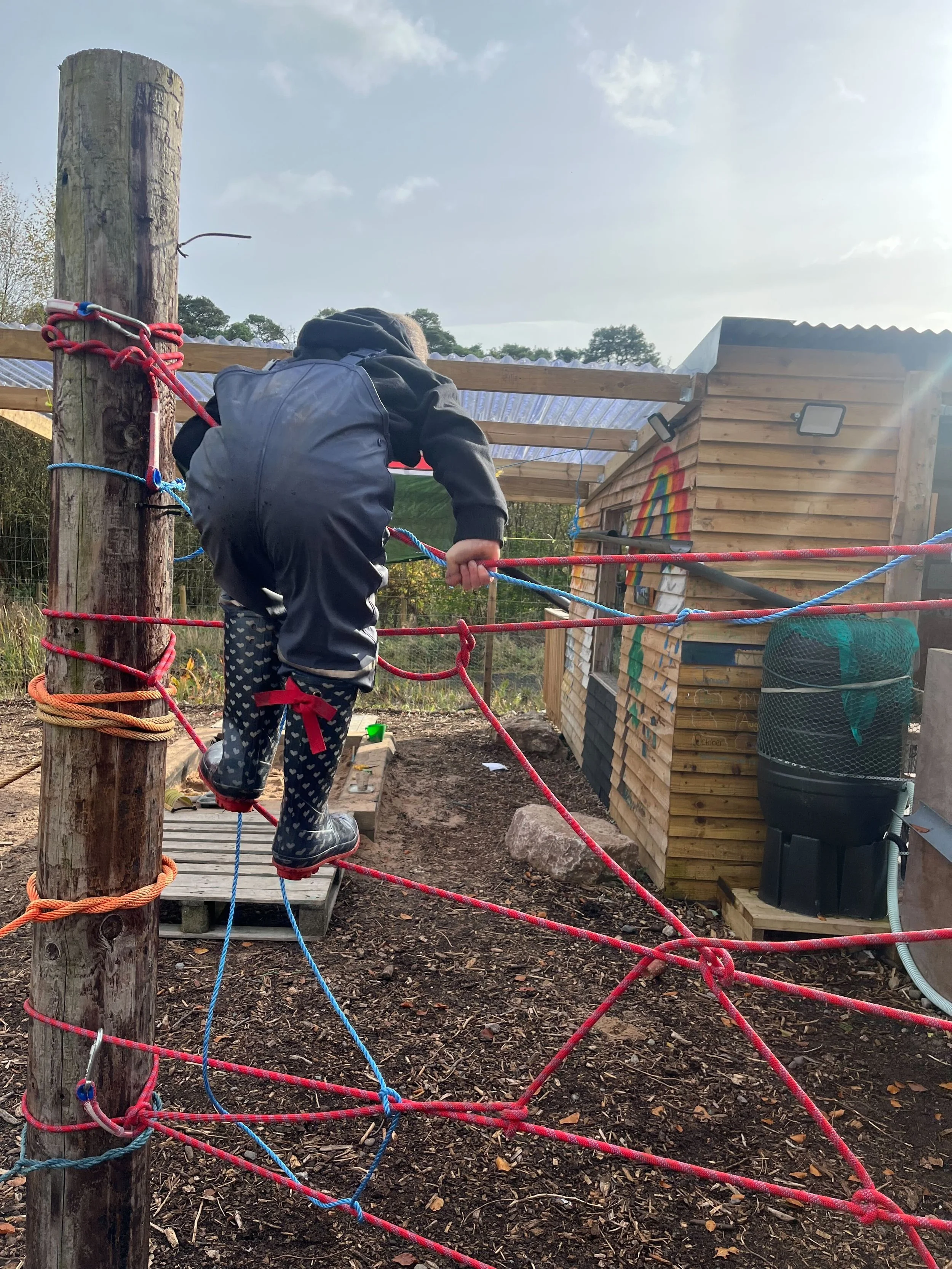 Child climbing along a rope obstacle course outdoors near a wooden building with a rainbow painted on the side, with trees and a cloudy sky in the background.